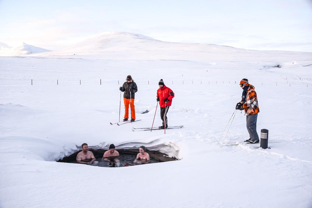 Three people bathing in a hot spring and three skiers standing next to them