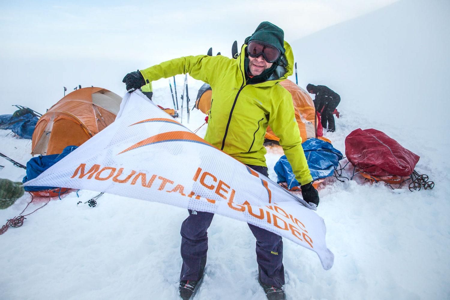 A person in a bright yellow jacket and black pants is holding a white banner with orange and black text, in a snowy environment with tents and gear in the background.