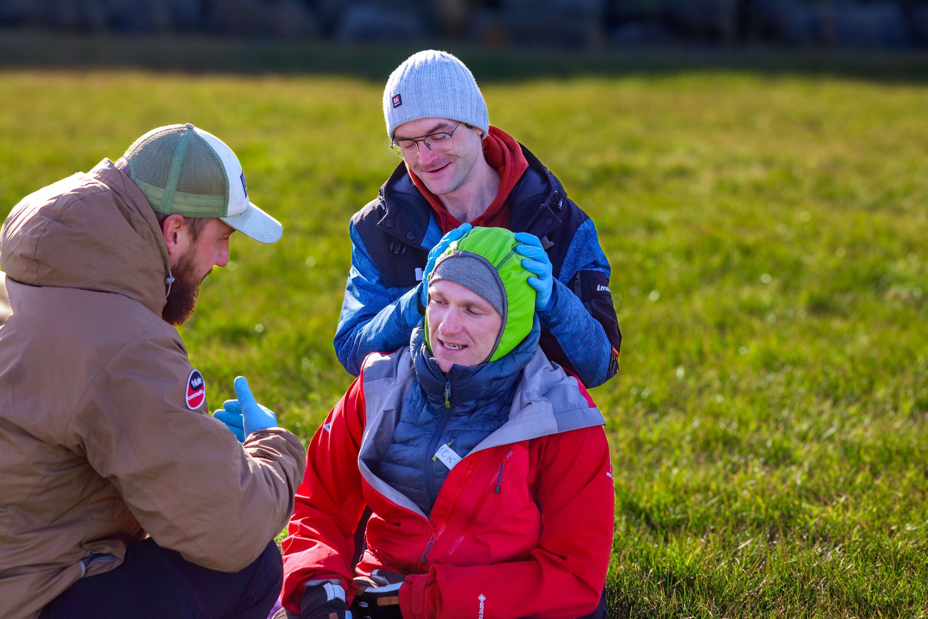 Three participants in a Wilderness First Responder course practice a head injury assessment on a simulated patient seated on grass, focusing on stabilizing and supporting the patient's head.