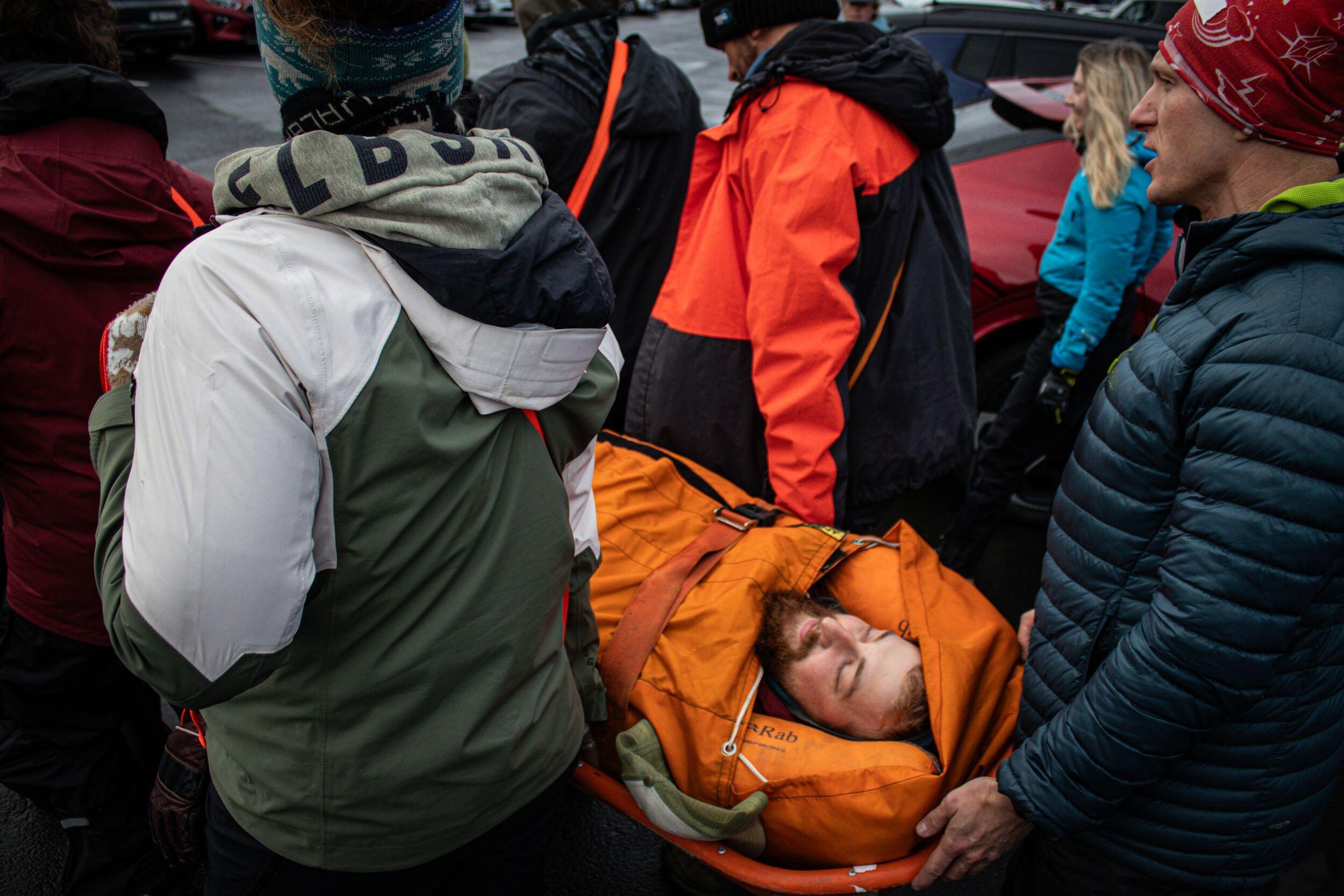 This image depicts a group of individuals participating in a simulated rescue, transporting a person on an orange stretcher who appears bundled in warm gear, with eyes closed, as part of a training exercise.