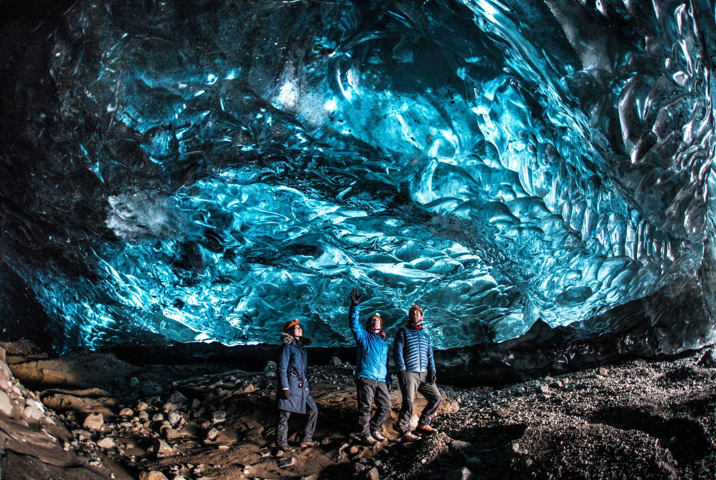 A person standing in an ice cave's interior with a translucent blue hue showcasing intricate patterns and textures resembling waves etched into the ice wall.