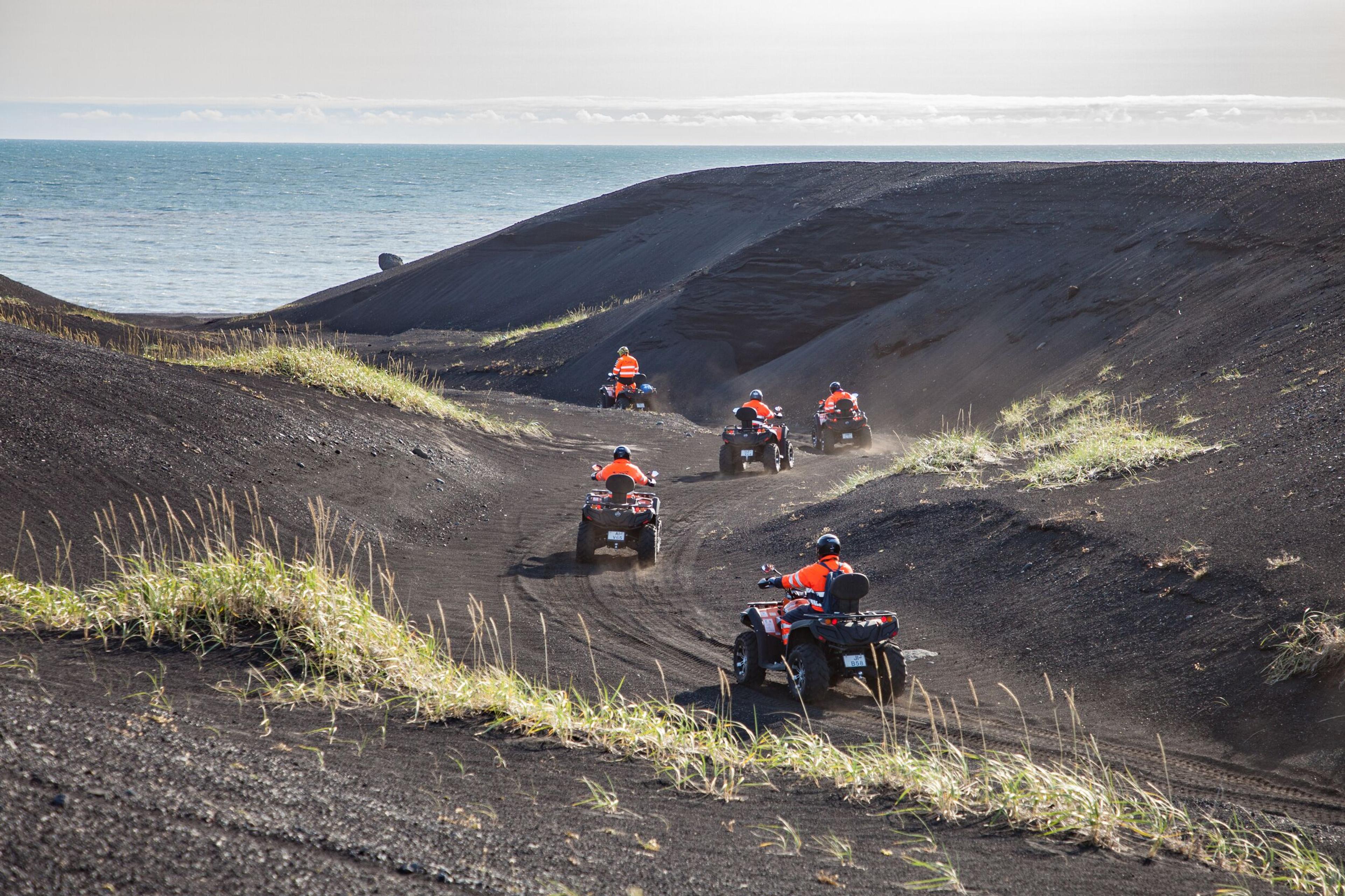  Express Tour with DC3 Plane Wreck Visit in Vik, Iceland: Several ATVs with riders in orange safety vests traverse a dark, volcanic landscape with the ocean visible in the distance.