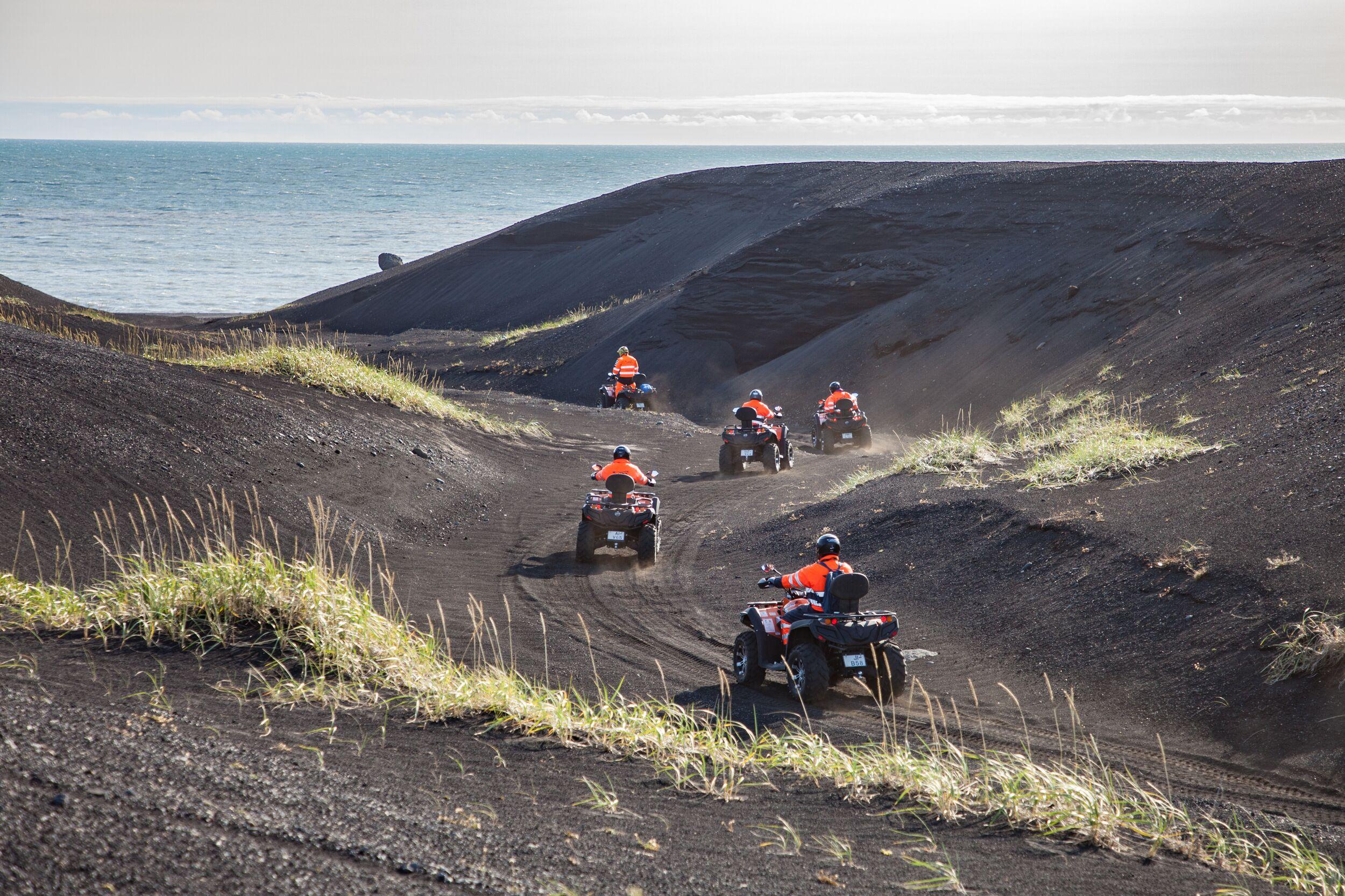 Express Tour with DC3 Plane Wreck Visit in Vik, Iceland: Several ATVs with riders in orange safety vests traverse a dark, volcanic landscape with the ocean visible in the distance.