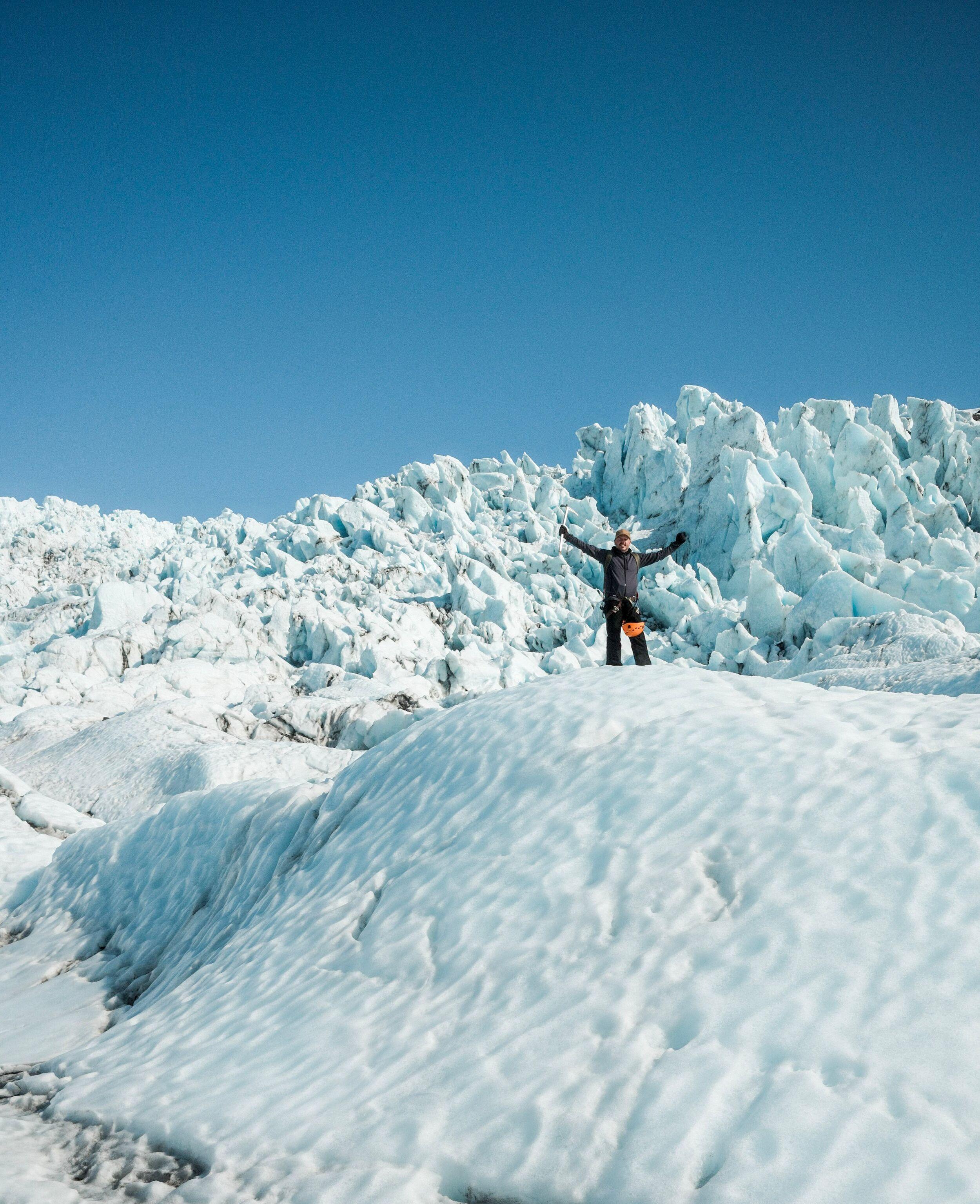 "A hiker raises an ice axe triumphantly while standing on a glacier ridge, dwarfed by jagged blue ice formations rising behind him