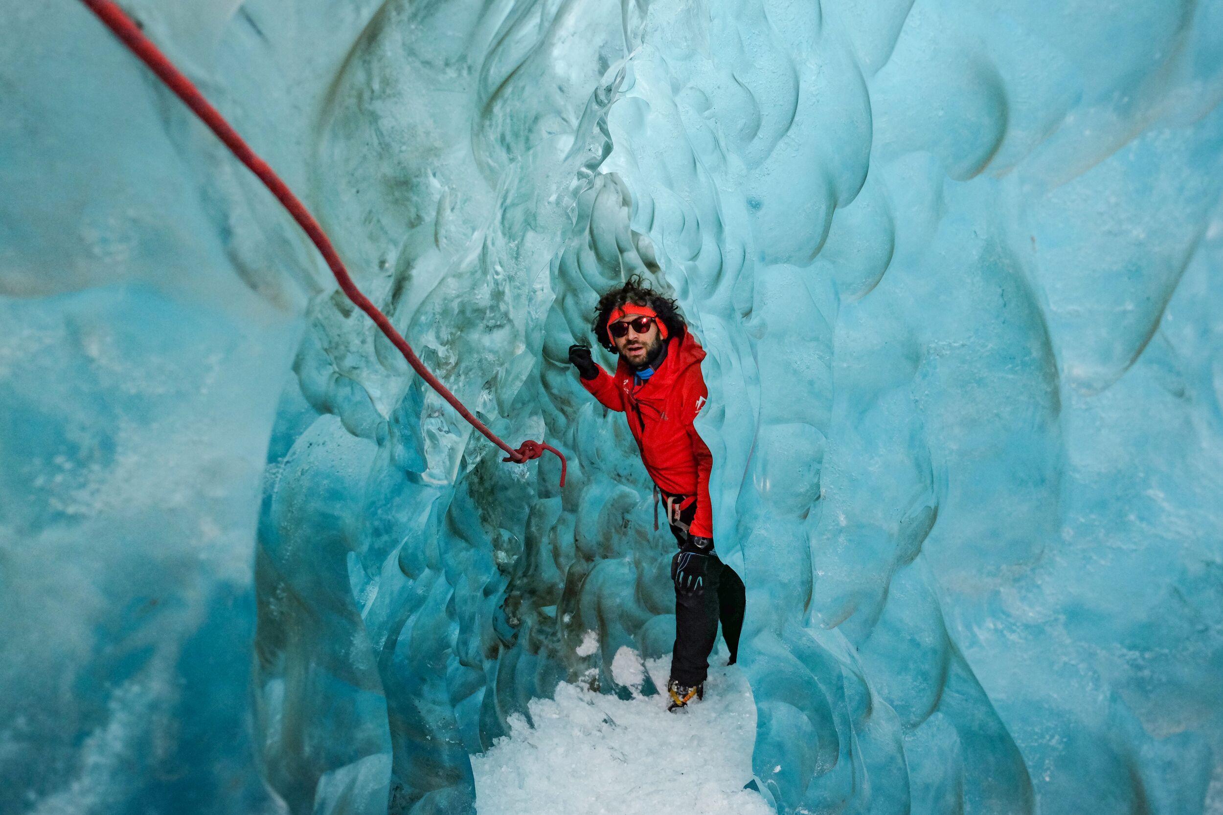 A person in a red jacket climbs a rope in a blue ice cave.