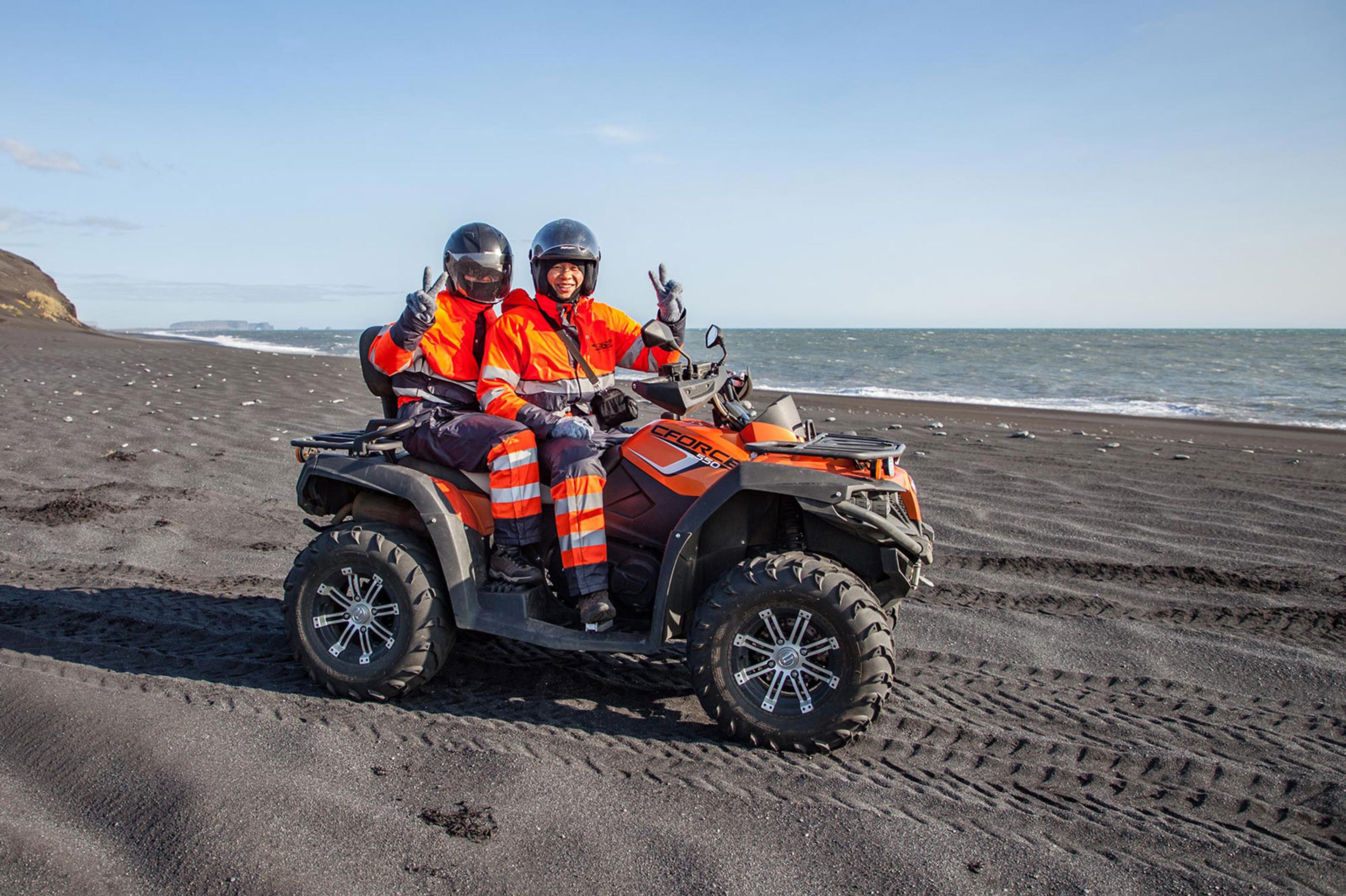 Two people sitting on an ATV quad bike happy and posing for the camera with the black sand and the ocean in the background