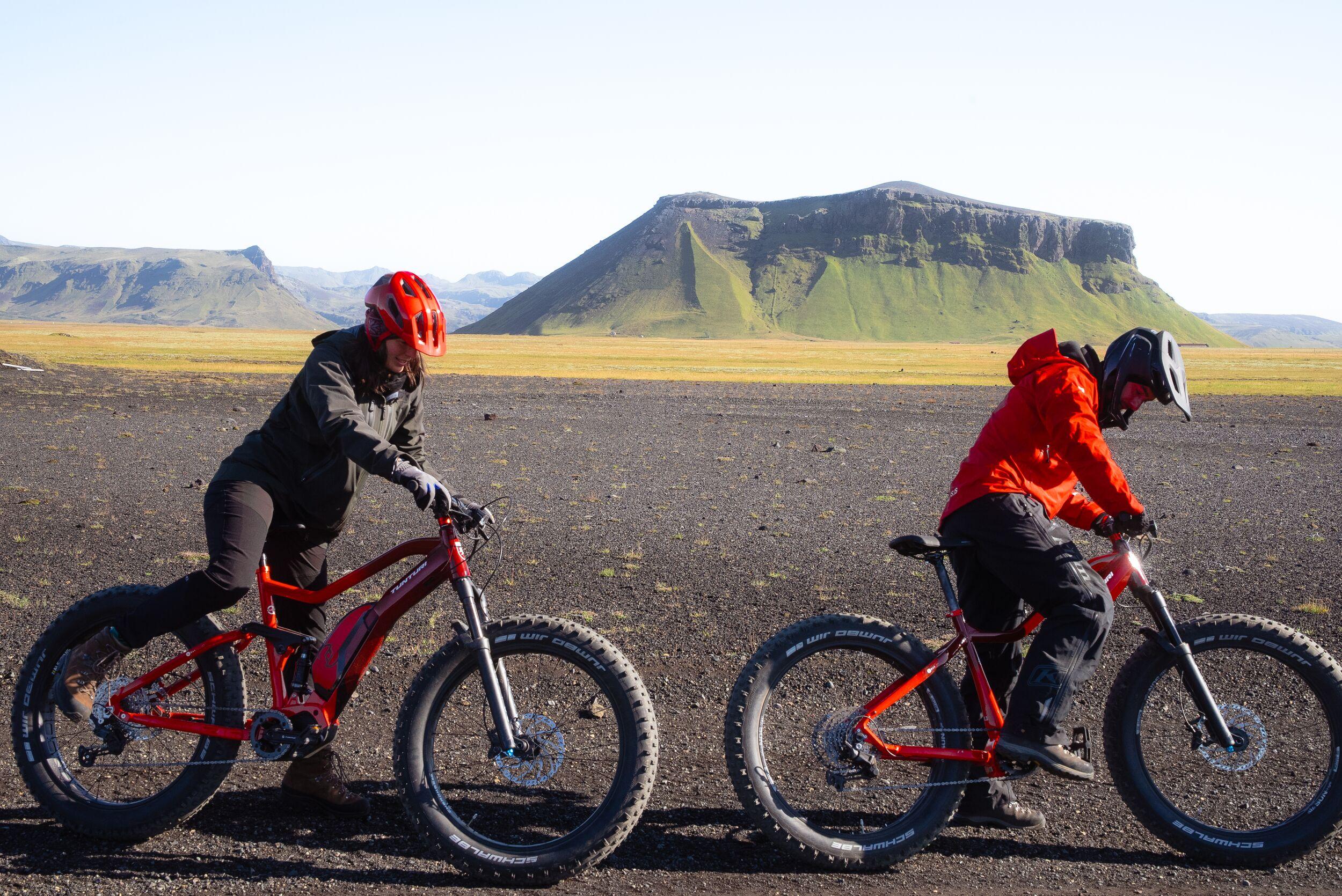 woman and a man on a bike on the black sand with a mountain in the back