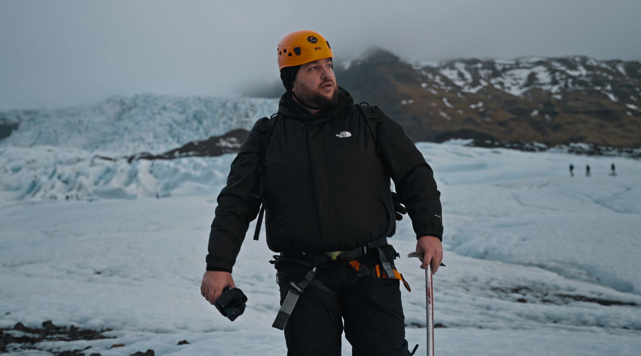 A hiker holding an ice axe stands inside a narrow glacier crevasse, surrounded by smooth walls of deep blue ice