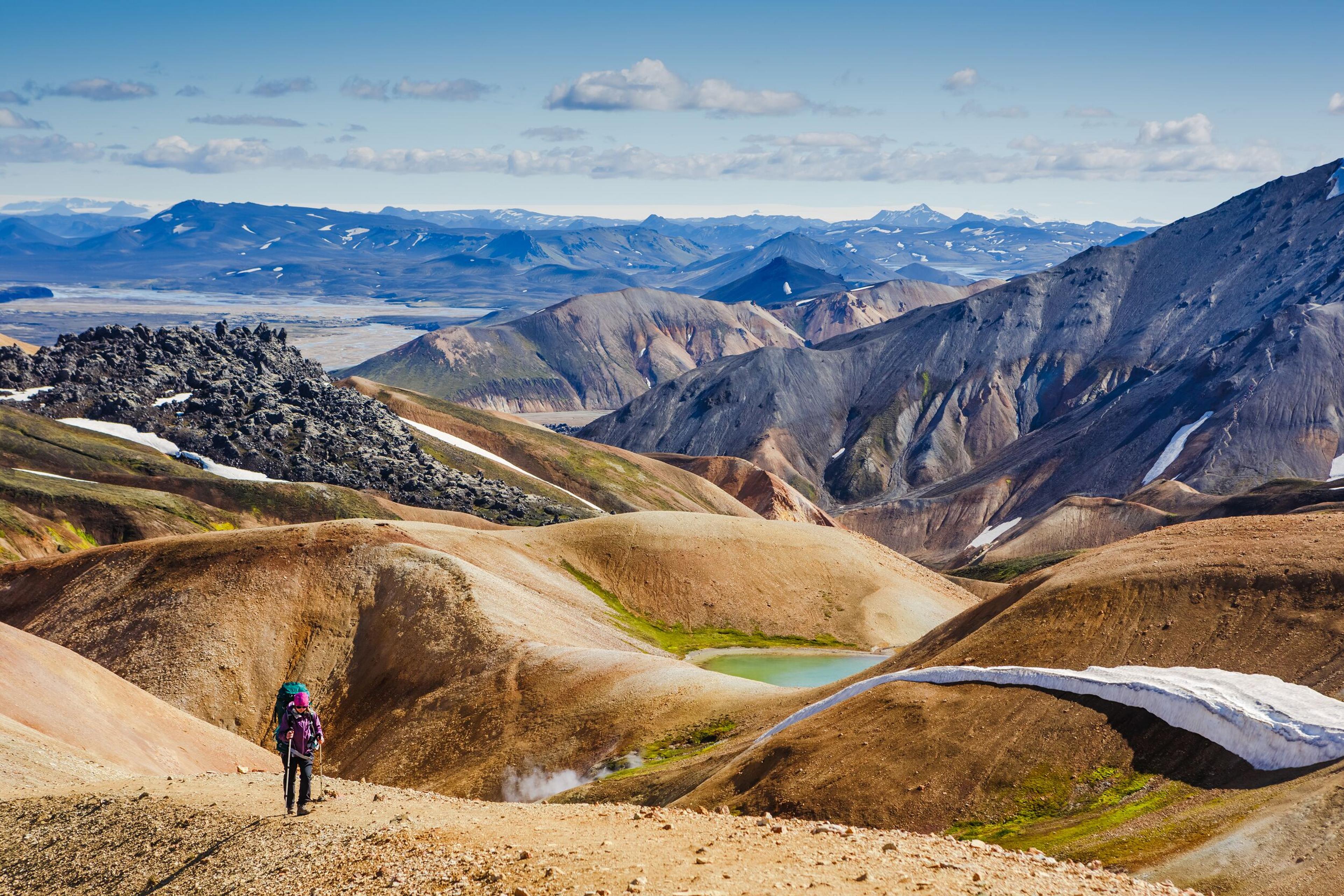 A hiker walks along a trail in Landmannalaugar, surrounded by colorful rhyolite mountains and a small turquoise lake nestled in the valley.