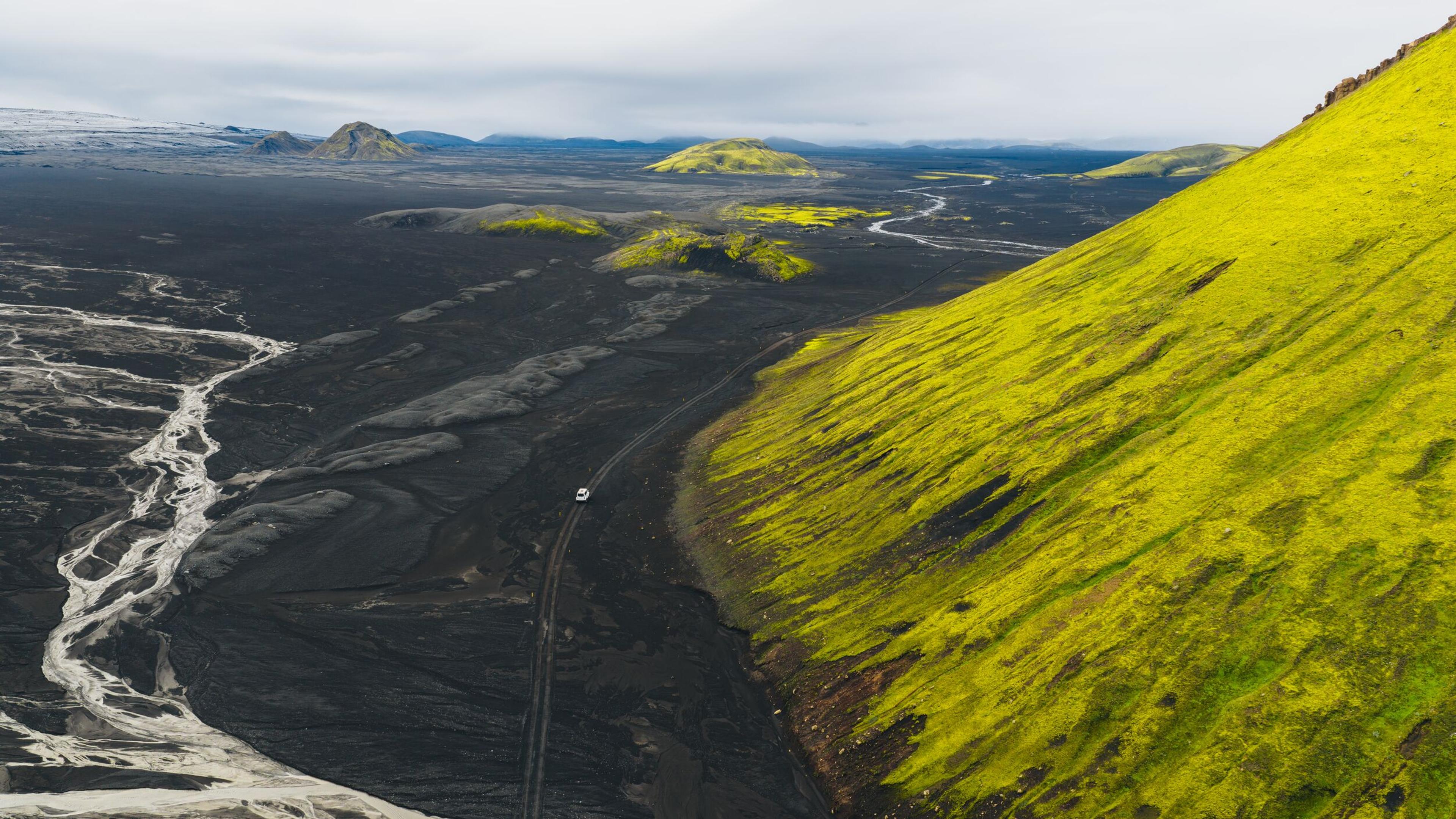 A single vehicle traveling on black sands with moss-covered bright green mountains all around