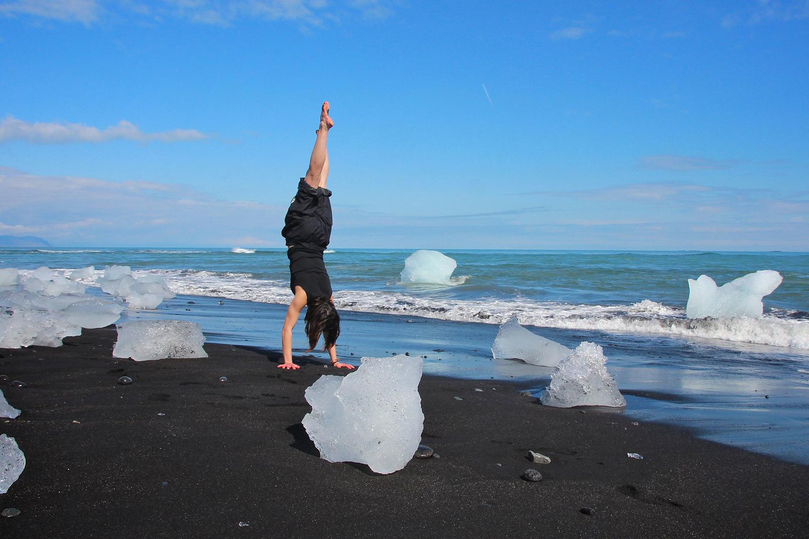 A Woman doing a hand stand in blacksand beach and icebergs around her.