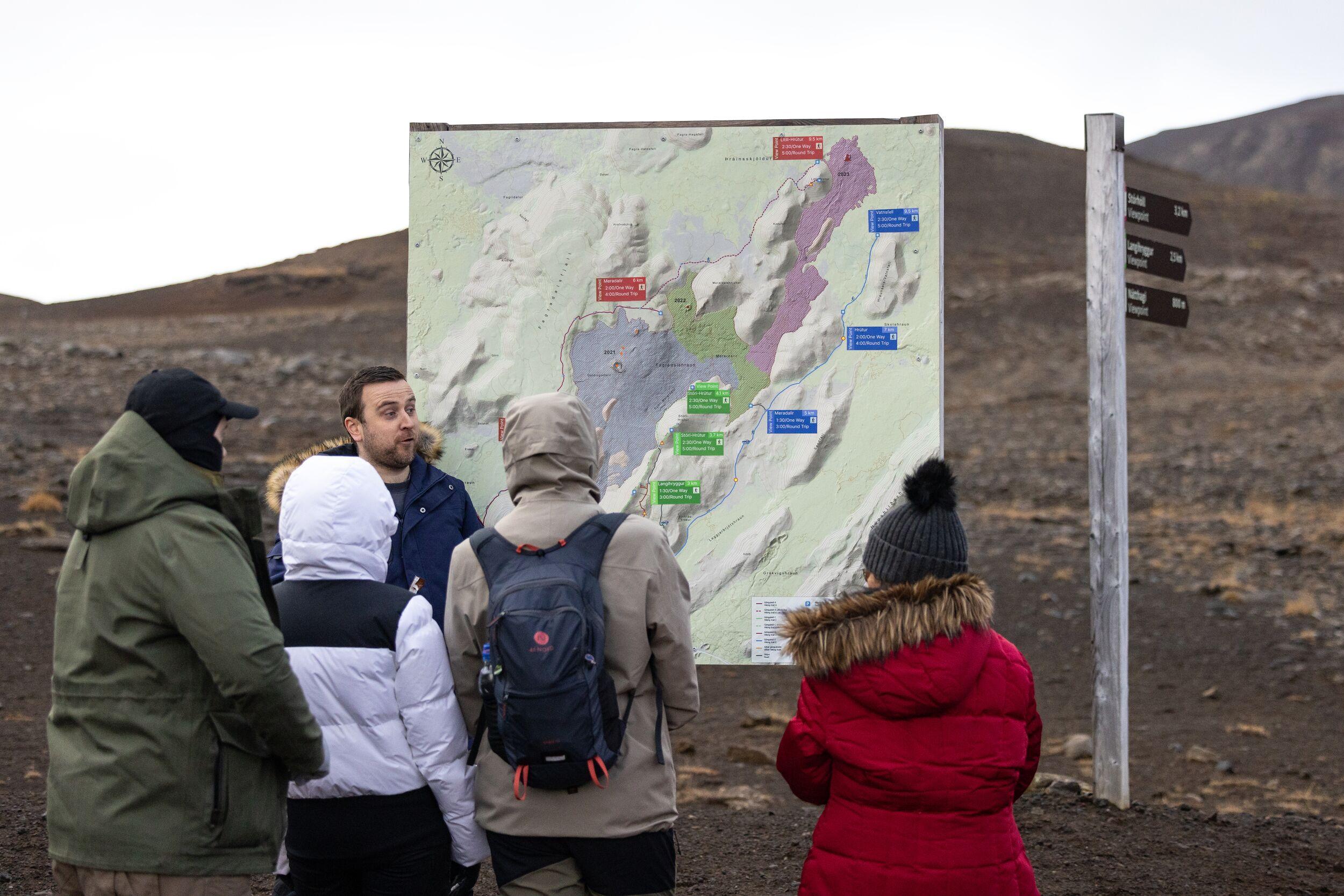 A group of people in winter clothing listens to a guide near a large map display, set against the barren volcanic landscape of the Reykjanes Peninsula.