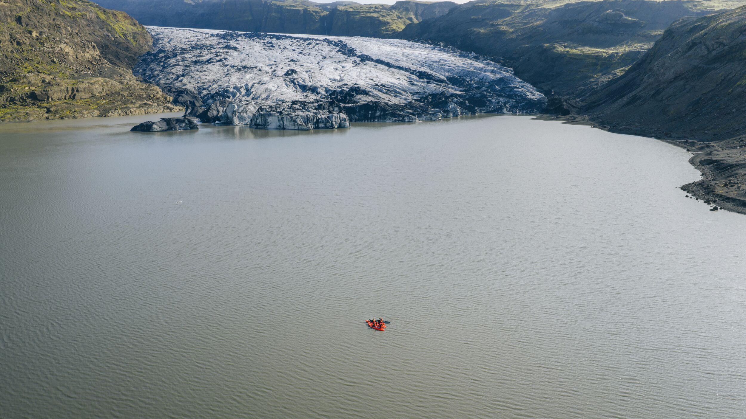 A glacier flows into a gray glacial lake surrounded by rugged, rocky mountains, with a small orange kayak near the center of the water.