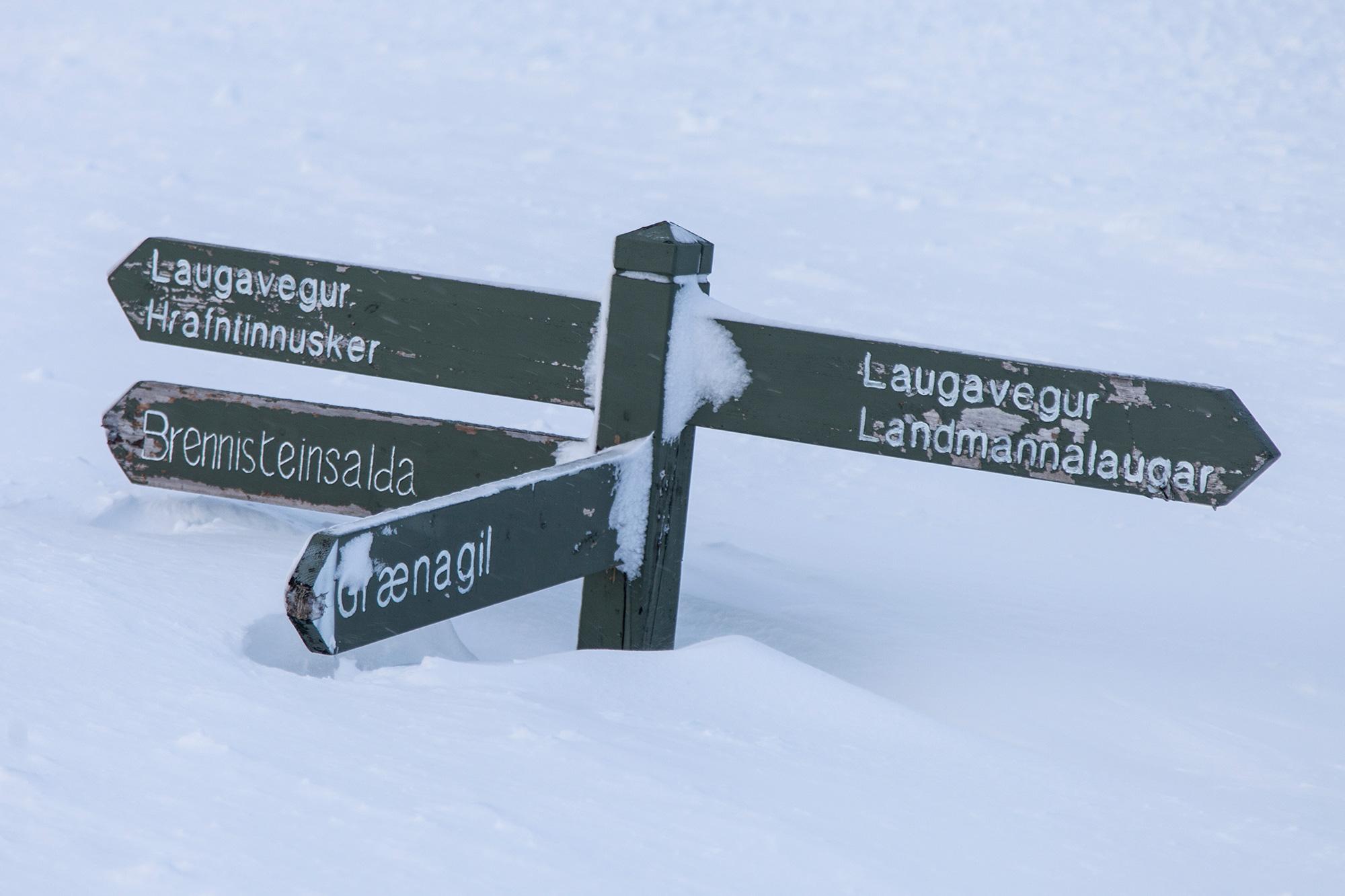 A directional signpost with multiple arrows is partially submerged in snow, with text indicating various hiking trail directions and distances.