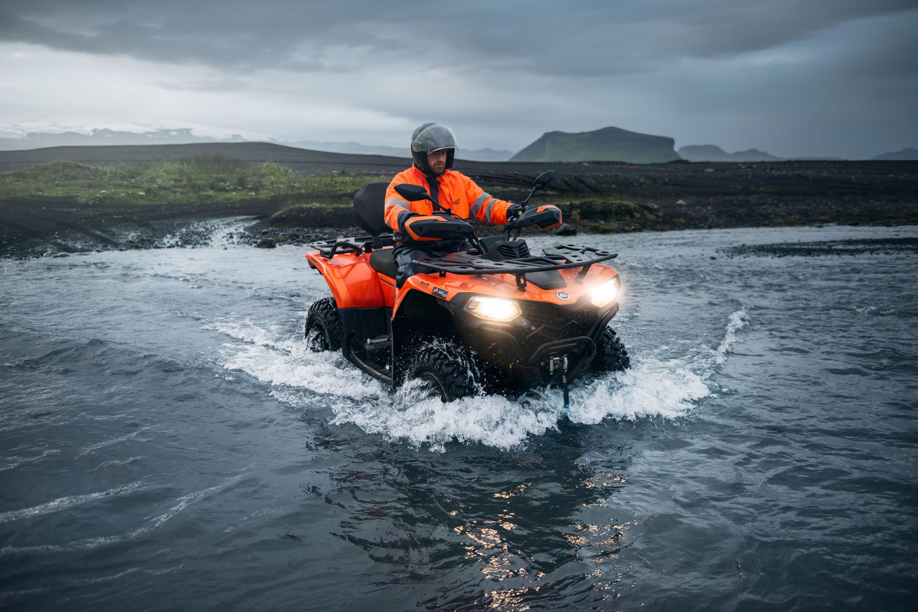 A rider in an orange waterproof jacket and helmet navigates an all-terrain vehicle (ATV) through a shallow river in Iceland.