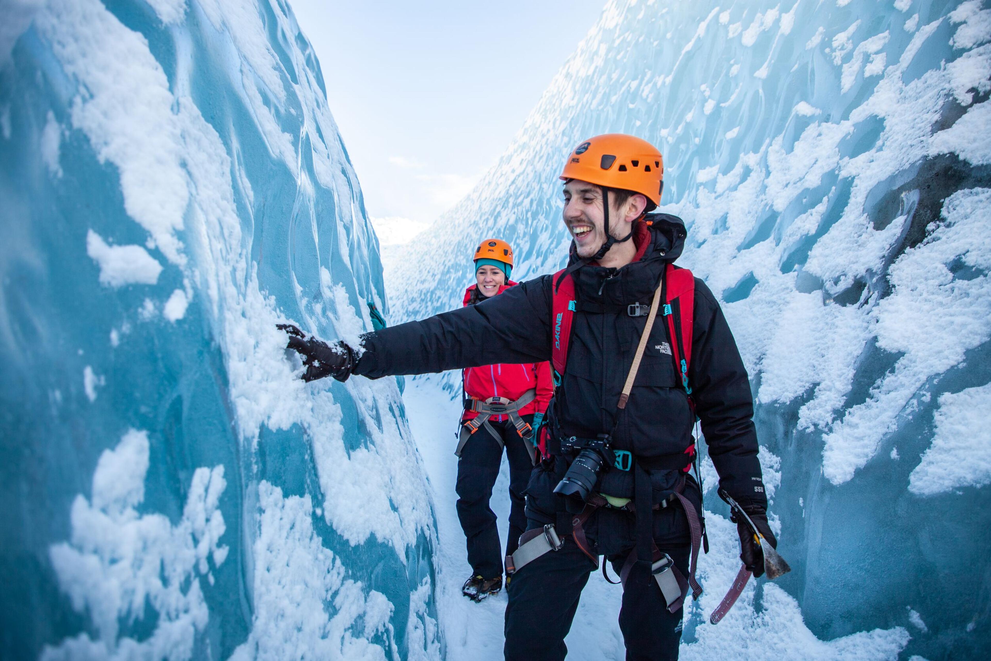 A smiling guide touches the wall of a deep blue glacier crevasse while explaining ice formations to a guest walking behind him