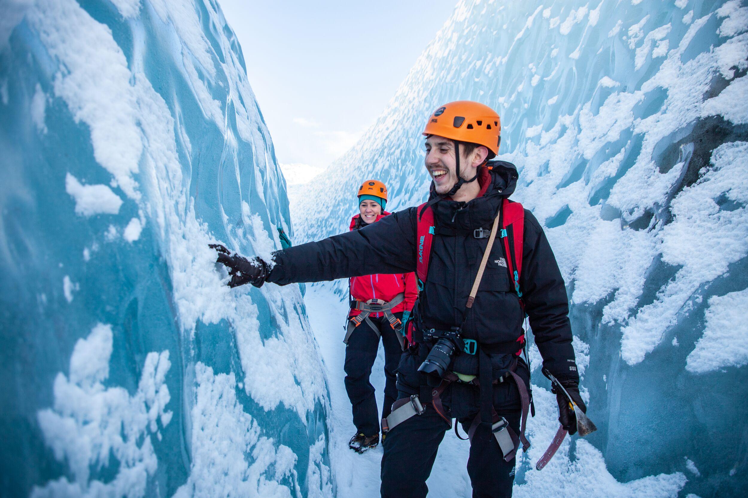 A smiling guide touches the wall of a deep blue glacier crevasse while explaining ice formations to a guest walking behind him