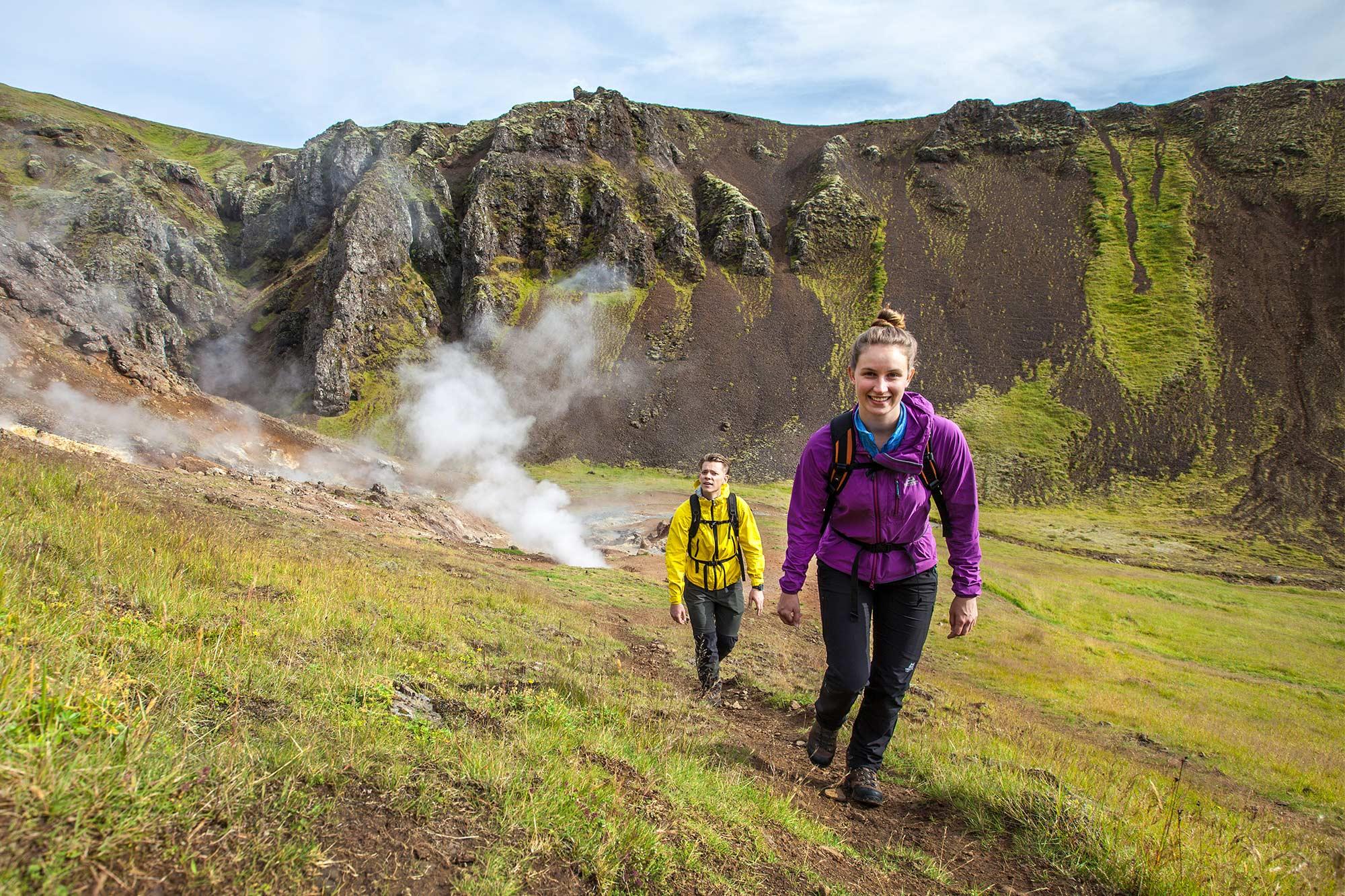 This image shows two hikers trekking along a scenic trail in Iceland, surrounded by rugged cliffs and geothermal steam vents. The lead hiker is wearing a purple jacket, followed by another in a yellow jacket, emphasizing the adventurous landscape and vibrant outdoor experience.
