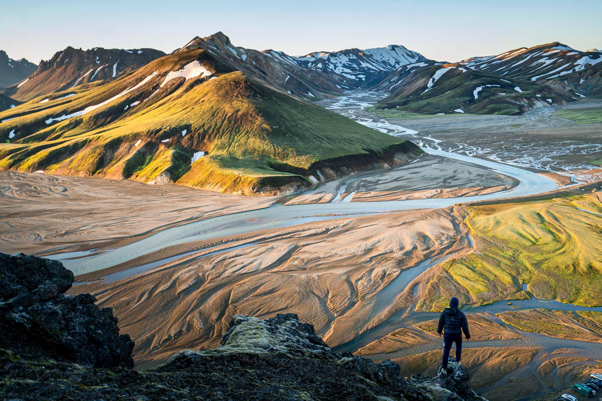 A hiker stands on a cliff overlooking the colorful, rugged terrain and river valleys of Landmannalaugar in Iceland.