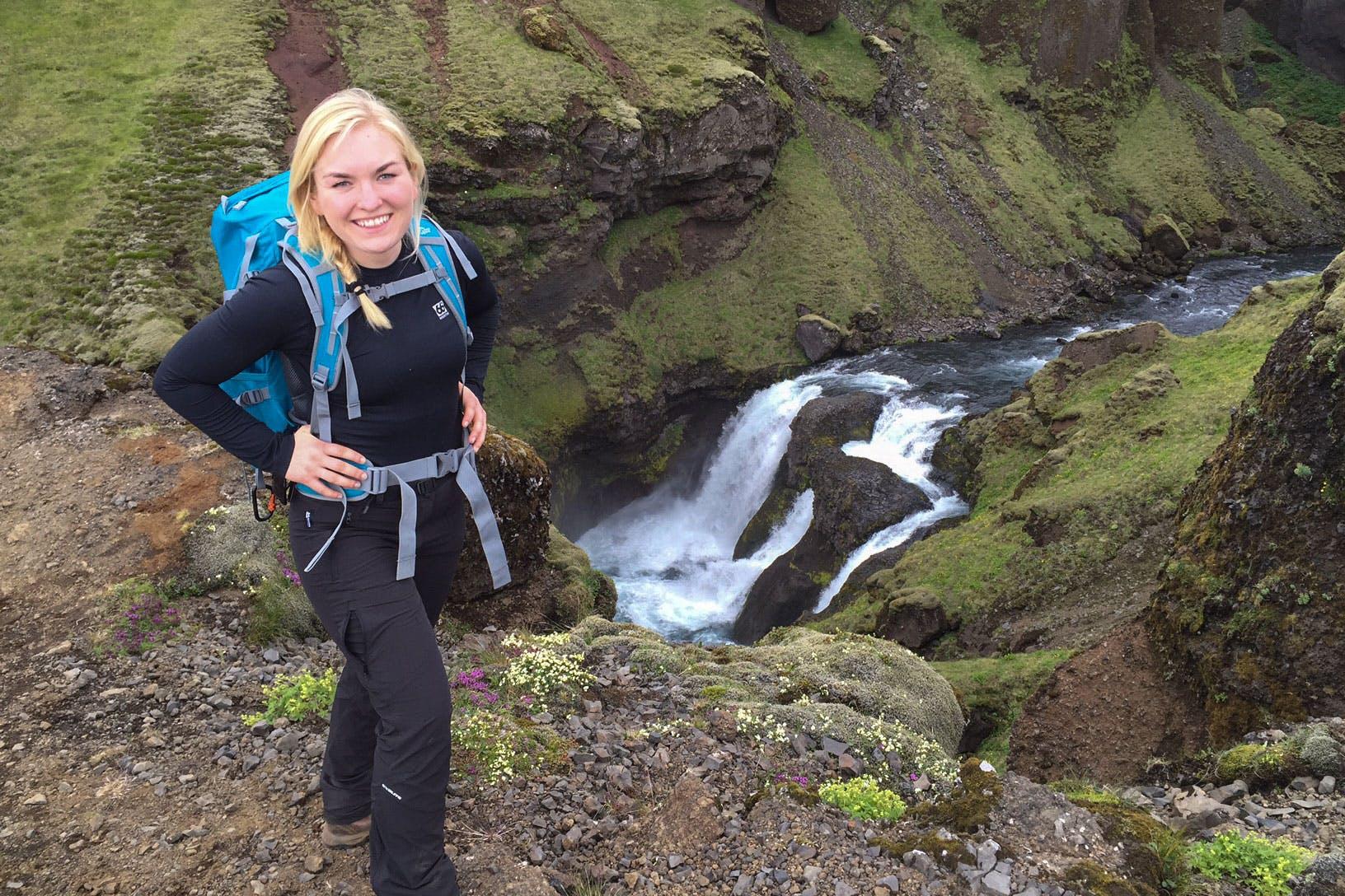 Woman smiling for picture with a backpack with river from Fimmvörðuháls trail in the background.