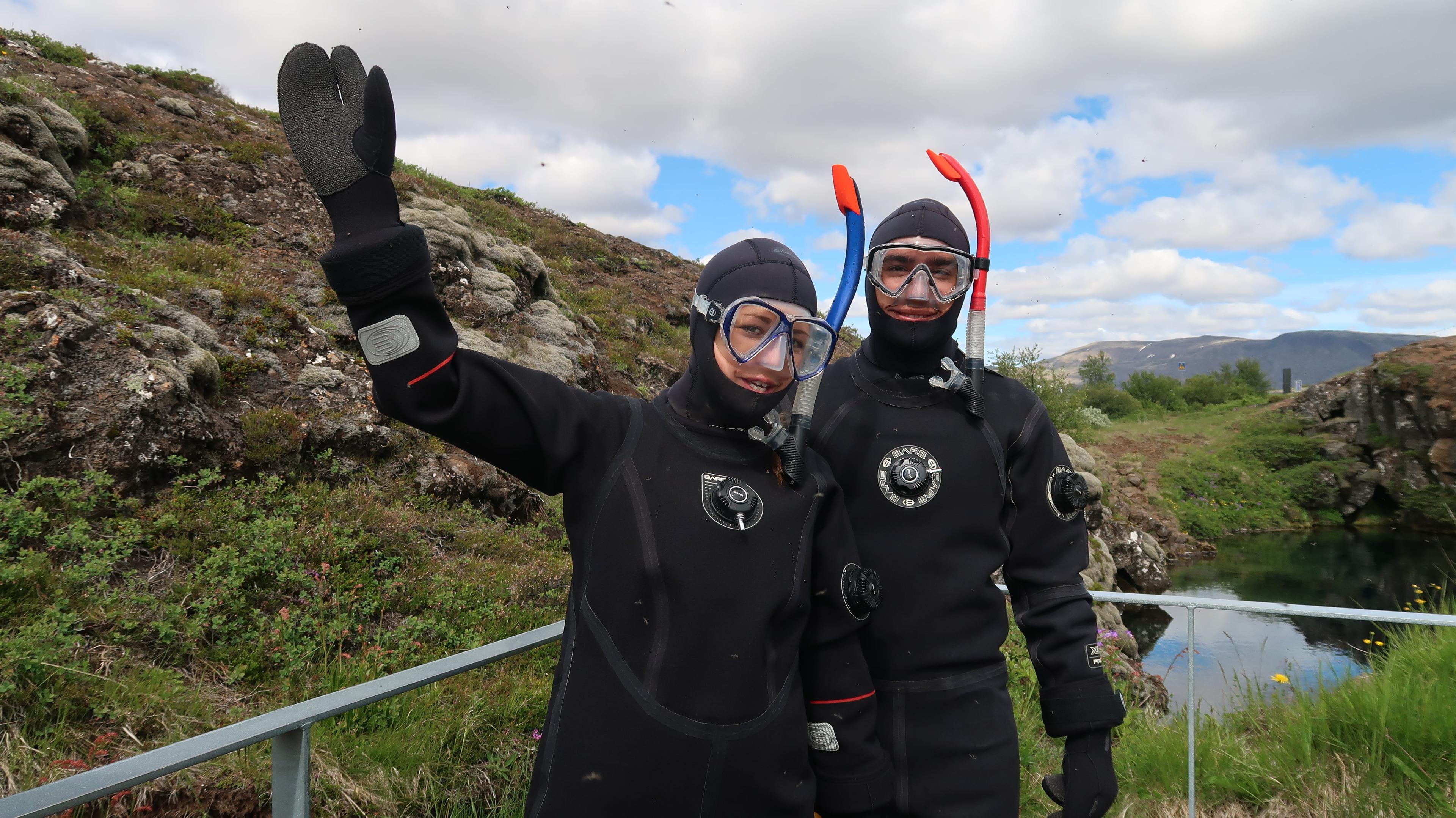 Two people dressed up in dry suits and snorkels waiving to the camera.