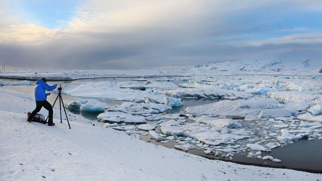 At the glacier lagoon in Iceland a man in a blue jacket is taking a photo of the icebergs floating on the water.