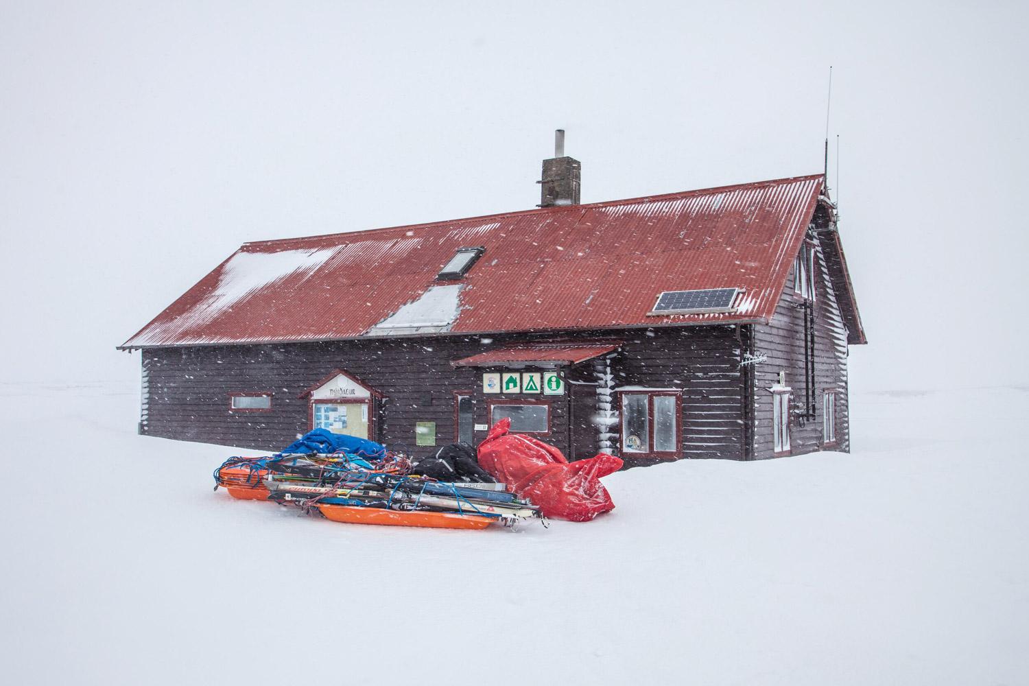 A wooden cabin with a snow-covered roof in a snowy landscape with colorful backpacks and winter gear placed in front of it.