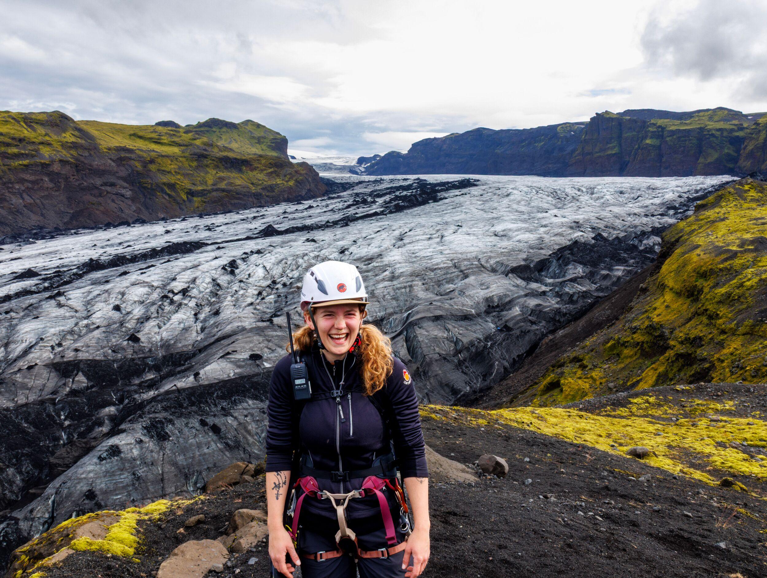 A smiling woman wearing a climbing helmet and harness stands on dark volcanic ground dotted with green moss, with a vast glacier and its crevassed ice stretching into the distance behind her.