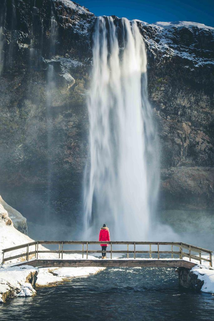 A woman standing on a wooden bridge in front of a waterfall in Iceland