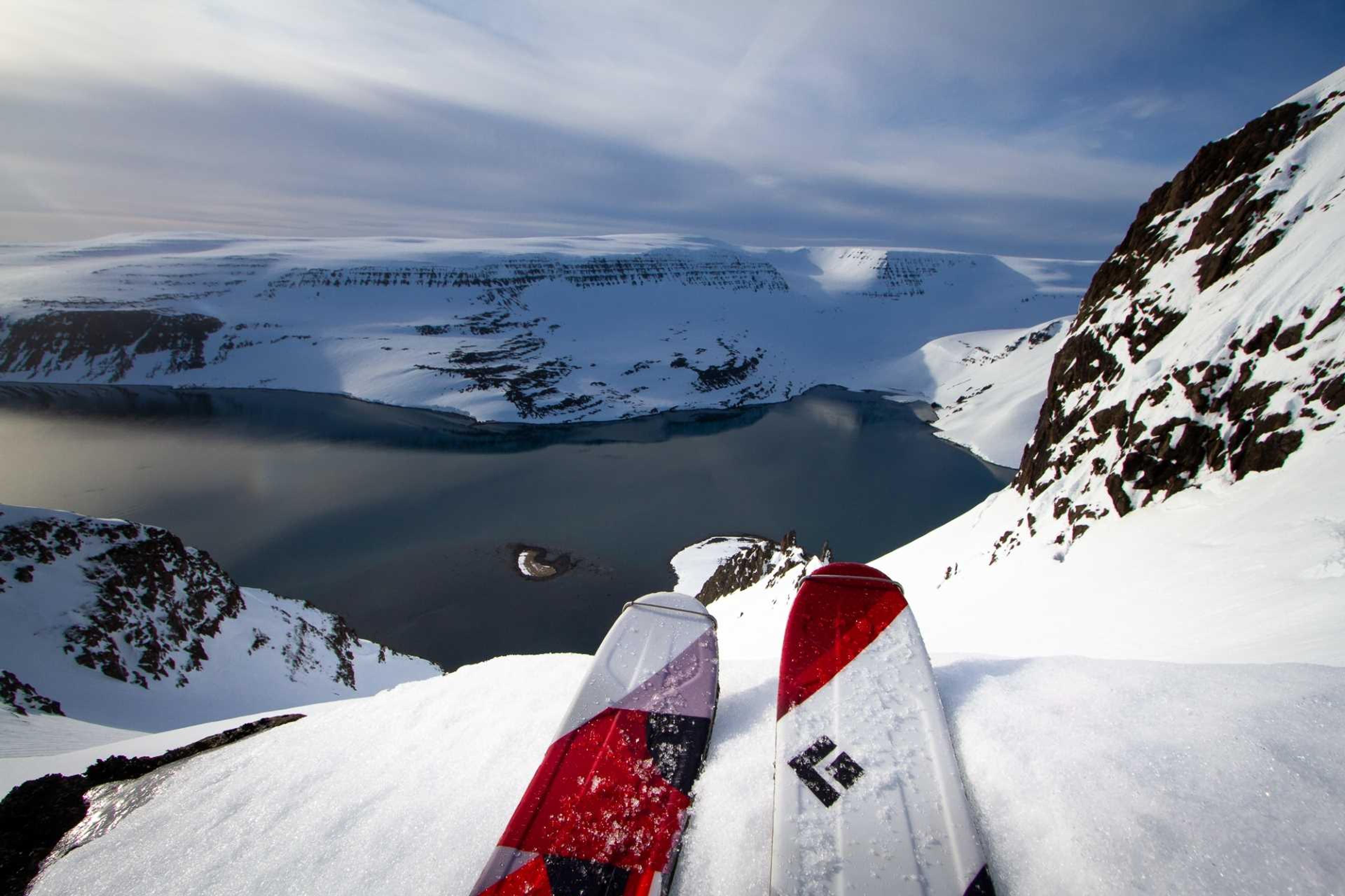 A pair of skis rests on a snowy ridge overlooking a stunning fjord surrounded by snow-covered mountains.