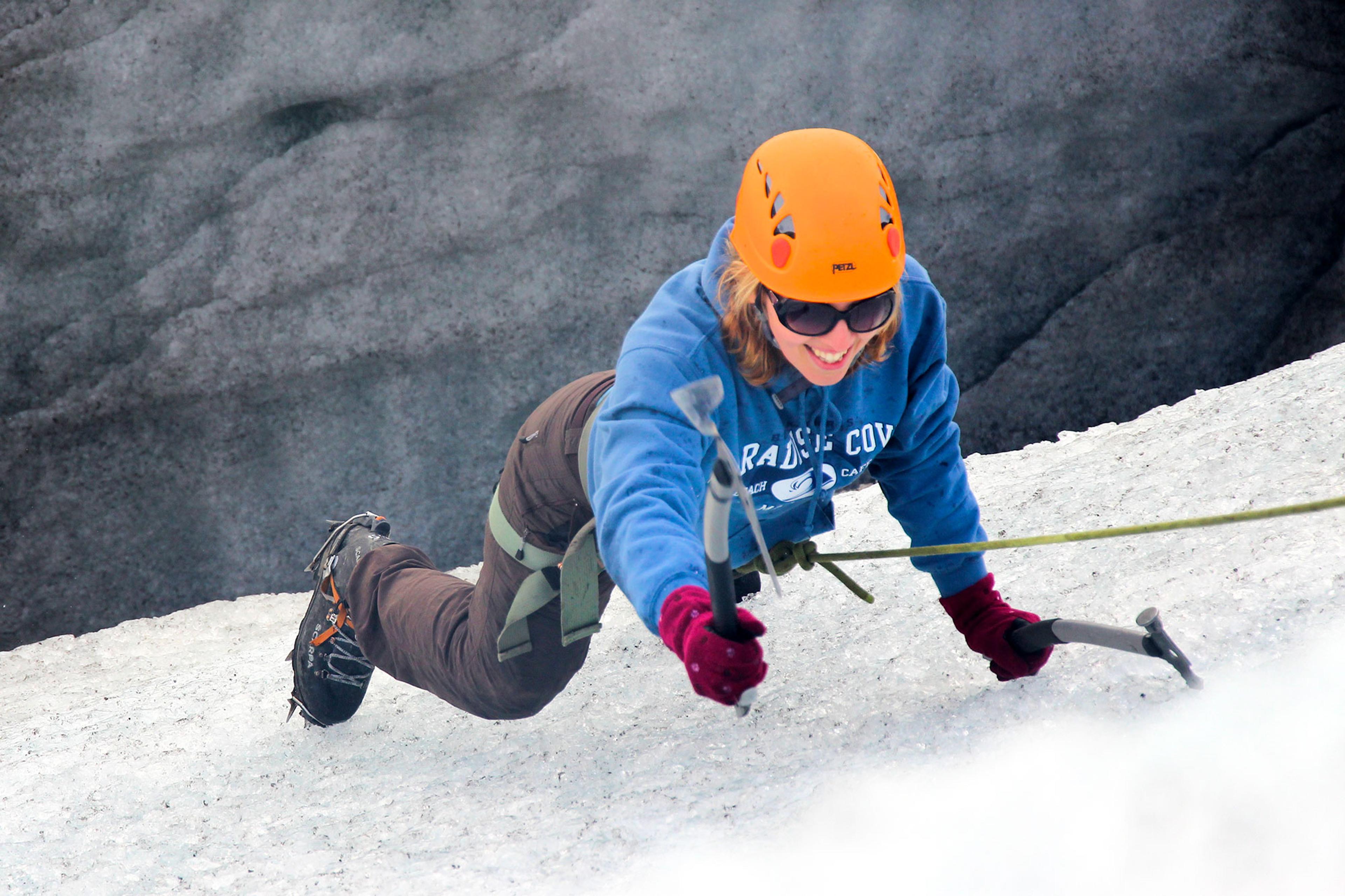 A smiling climber wearing a blue sweatshirt and orange helmet ascends an icy slope with the help of a rope and axes.