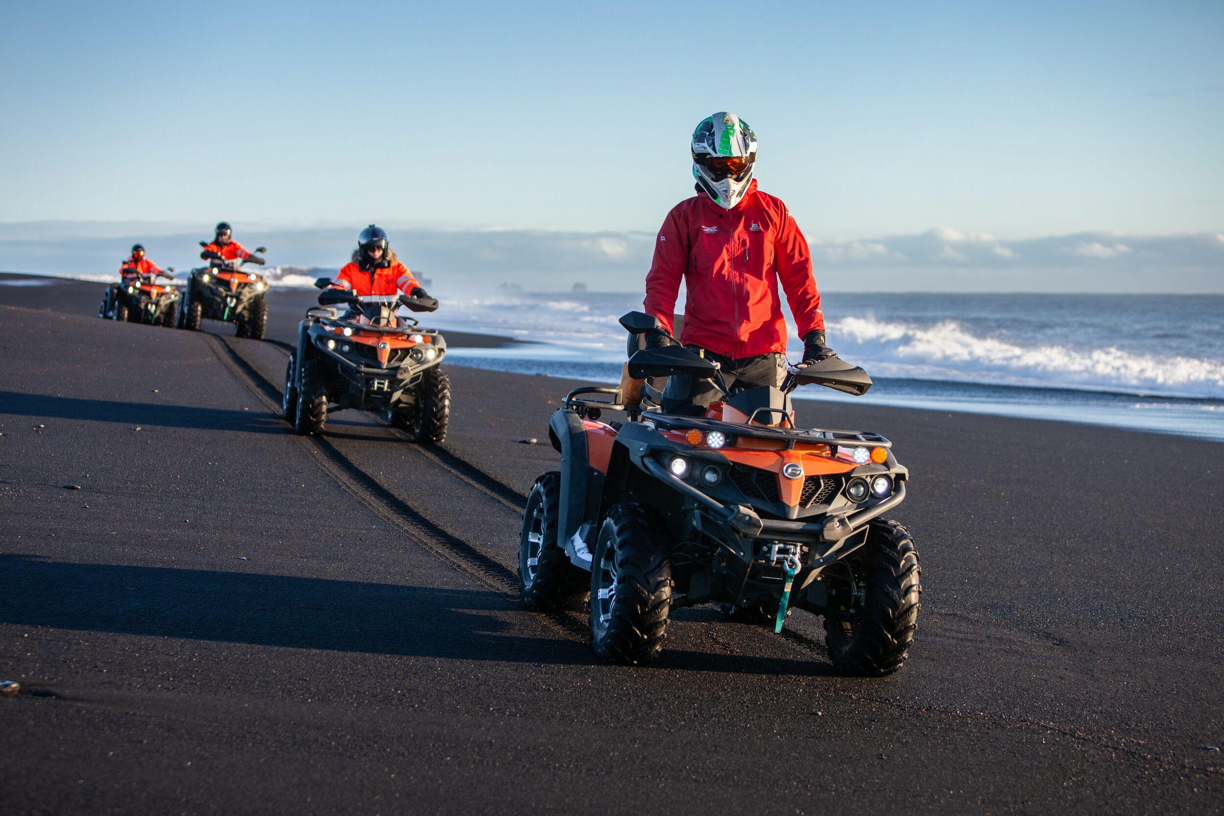 A group of ATV riders in protective gear cruise along Iceland's black sand beach, leaving deep tracks near the shoreline under a clear blue sky.