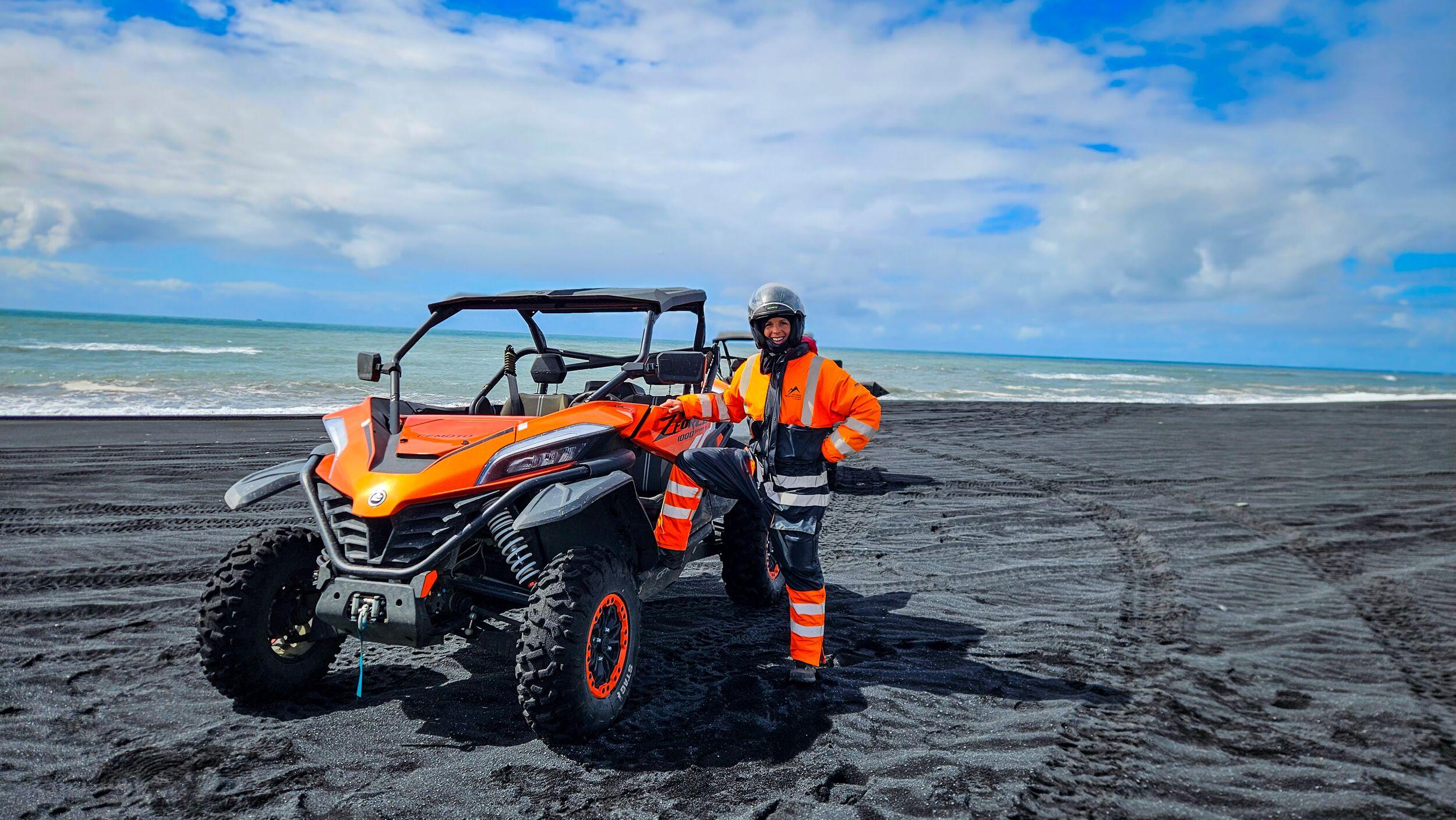 Person in bright orange gear standing next to an orange two-seater buggy on a black sand beach in Iceland, with the ocean and cloudy blue sky in the background.