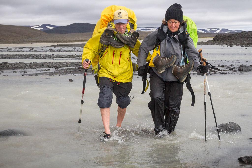 Two hikers in bright yellow rain gear crossing a shallow river, braving the elements while hiking in Iceland.