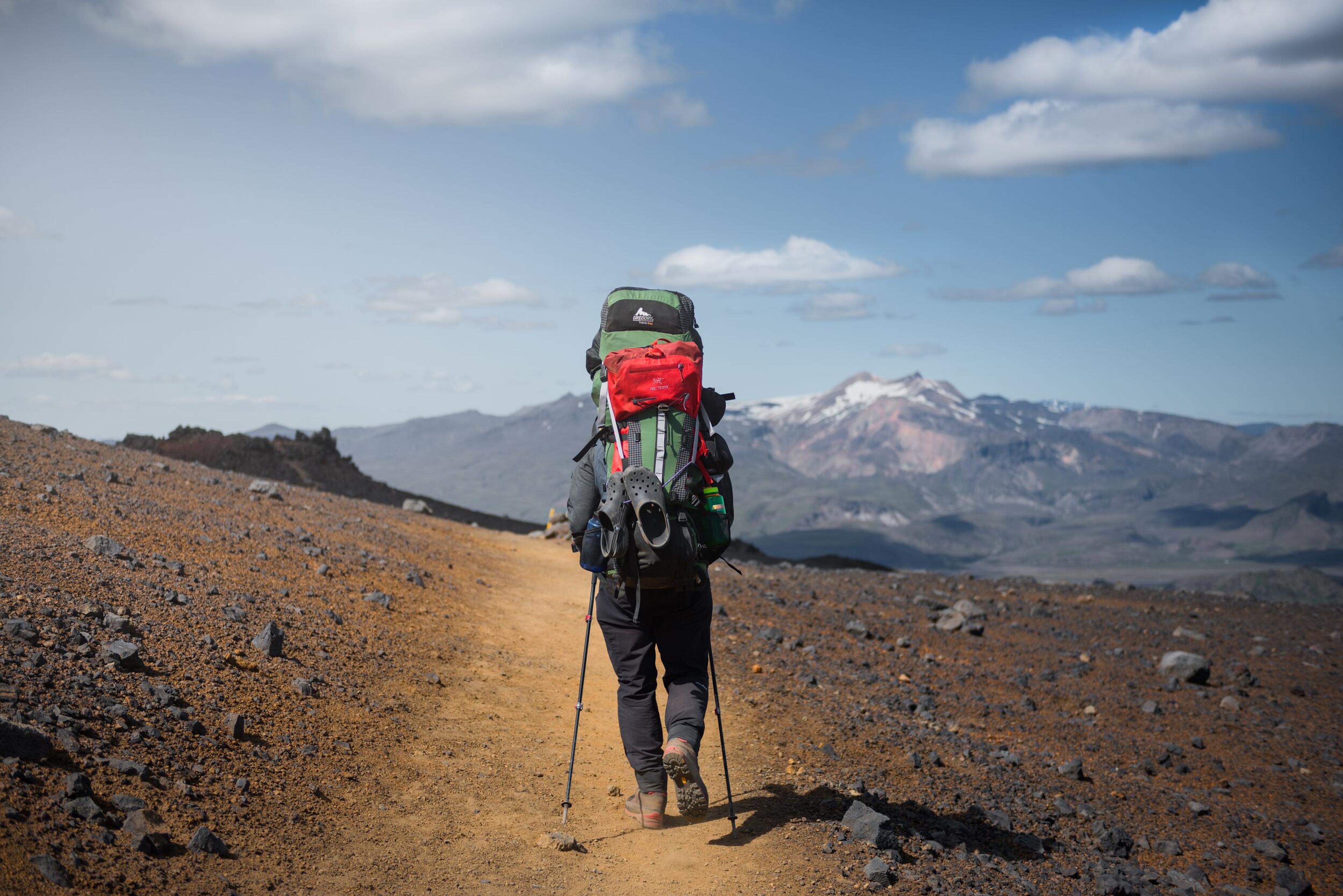 A hiker carrying a large red backpack and trekking poles walks along a dirt trail through barren, volcanic terrain in the Icelandic Highlands. Snow-capped mountains and a clear blue sky stretch out in the distance, highlighting the rugged beauty of the landscape.