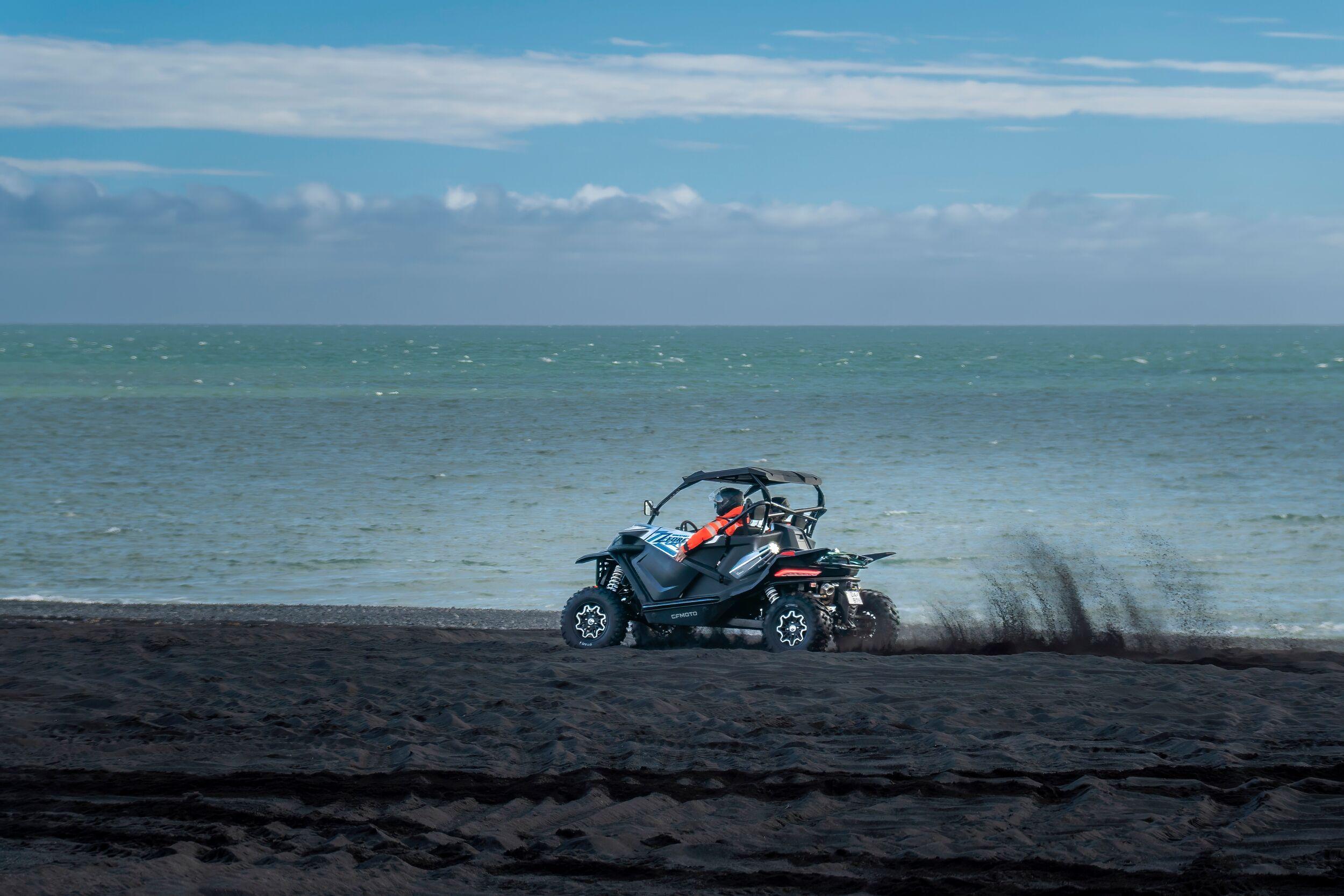 An off-road buggy kicks up black sand as it speeds along a beach with the ocean stretching out under a partly cloudy sky, capturing the thrill of coastal exploration in Iceland.
