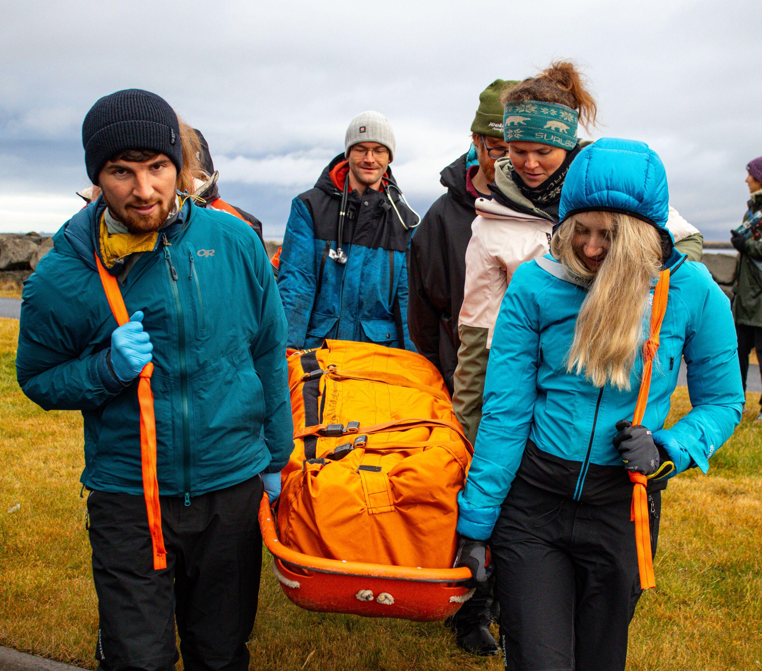 a group of individuals participating in a simulated rescue, transporting a person on an orange stretcher who appears bundled in warm gear, with eyes closed, as part of a training exercise.