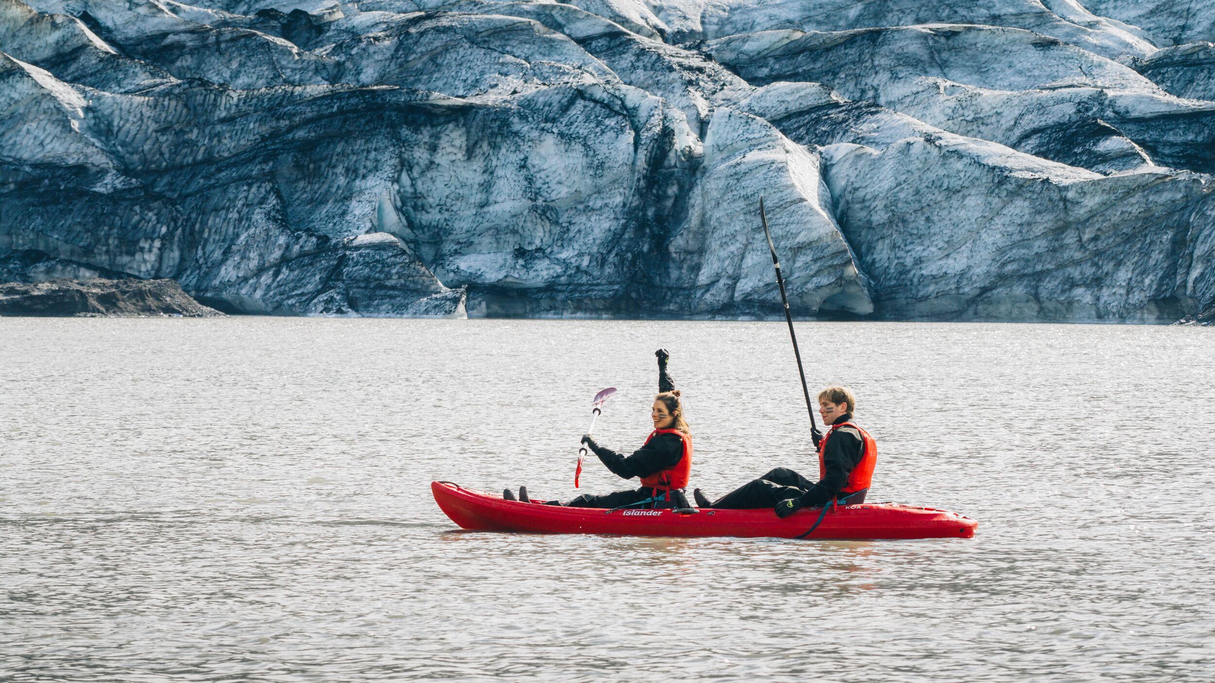 Two people in a red tandem kayak paddle across a glacial lake, with a massive blue-white glacier rising directly behind them.