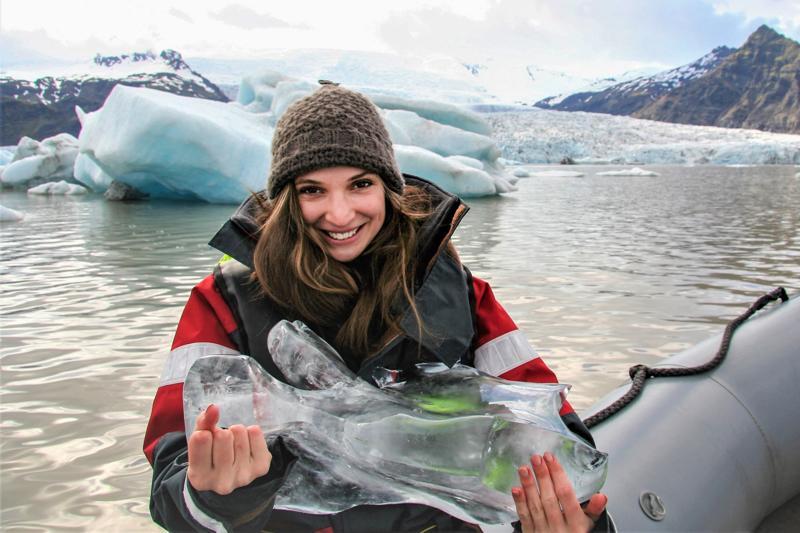 A smiling woman on an inflatable boat holds a chunk of clear glacier ice, with icebergs floating in a glacial lagoon and mountains behind her