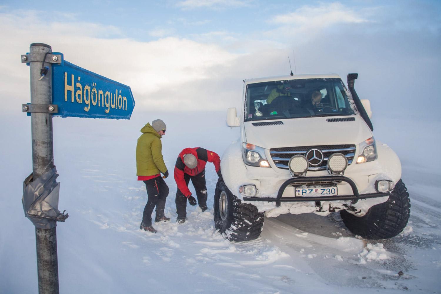 A four-wheel-drive vehicle is stuck in the snow near a signpost" with two people attempting to dig it out.