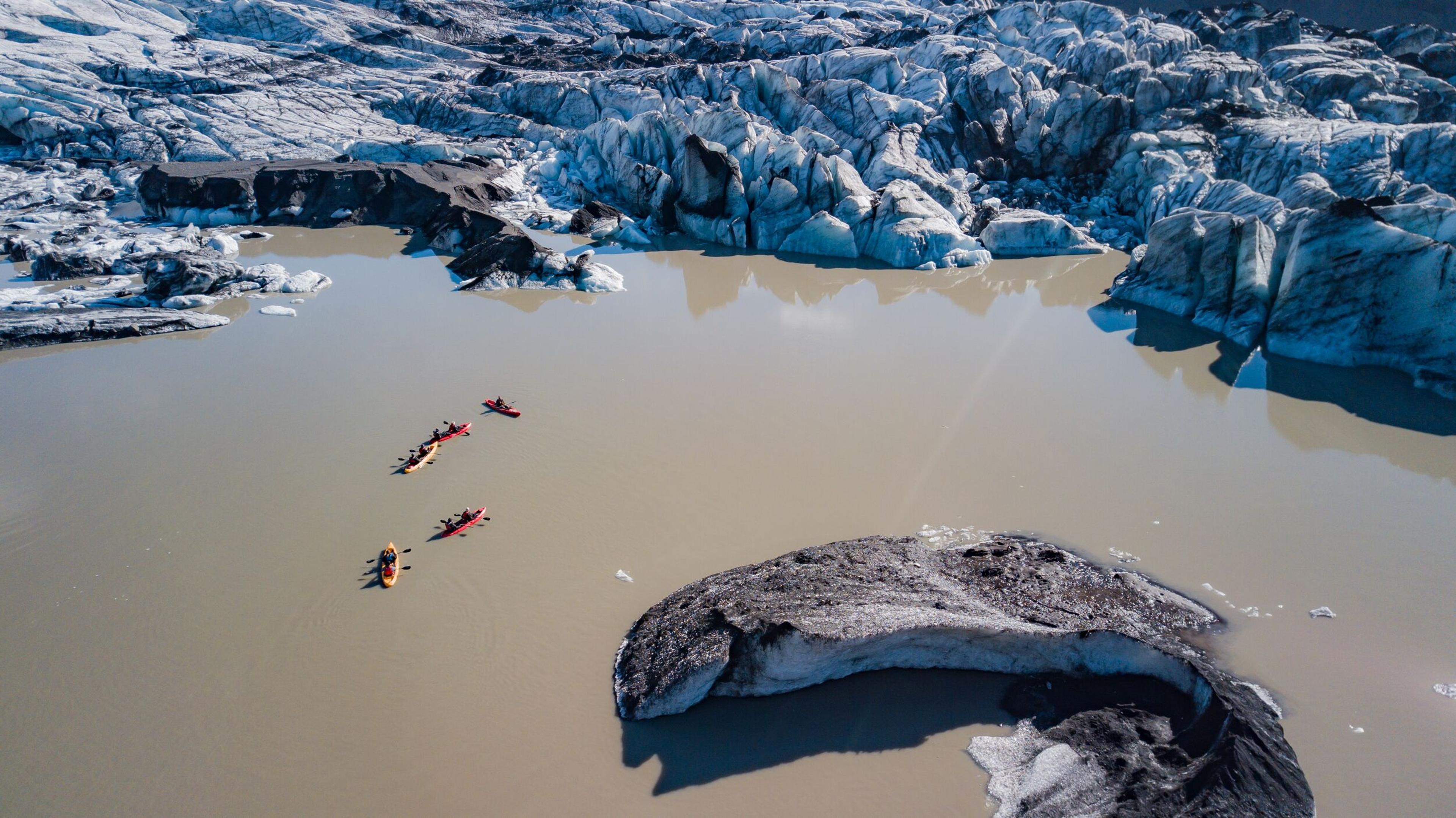 Aerial view of five colorful kayaks paddling across a milky glacial lake, surrounded by dramatic blue-white glacier ice and dark, debris-covered ice formations.