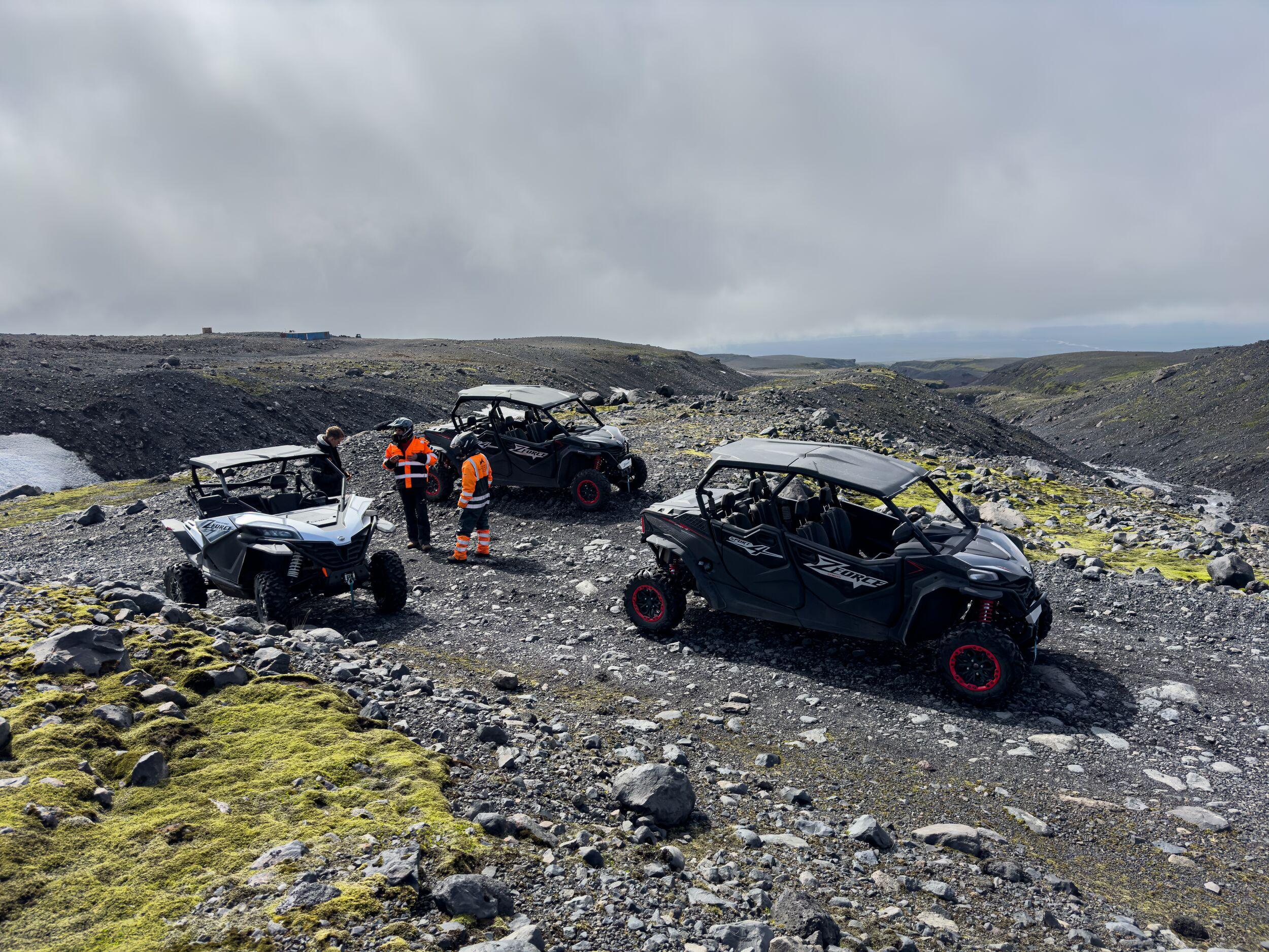 "Three off-road buggies are parked on a rugged Icelandic landscape as riders in bright orange gear take a break under a cloudy sky."