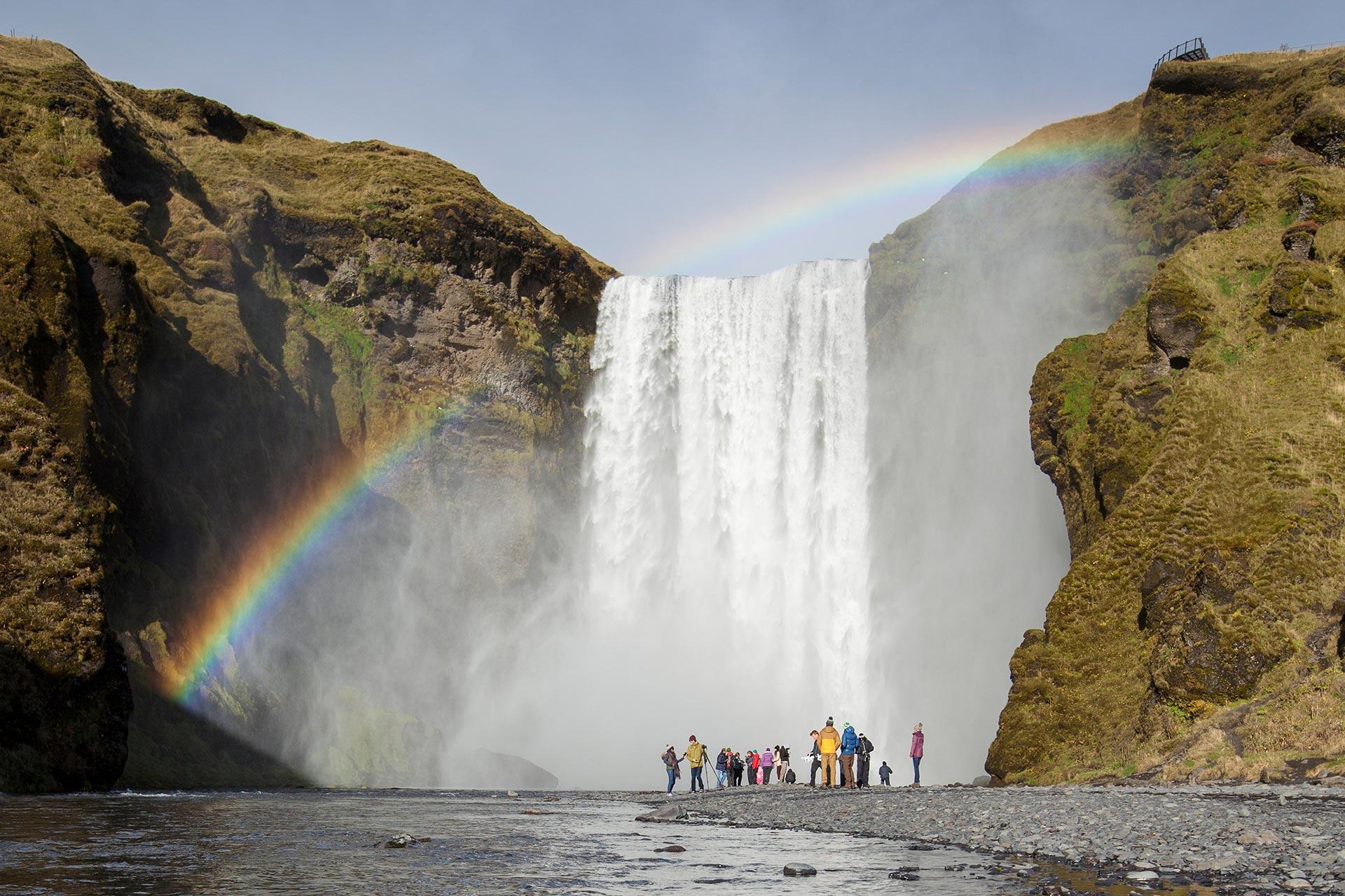People admiring Skogafoss (Skógafoss) waterfall in Iceland with rainbow shining across the waterfall