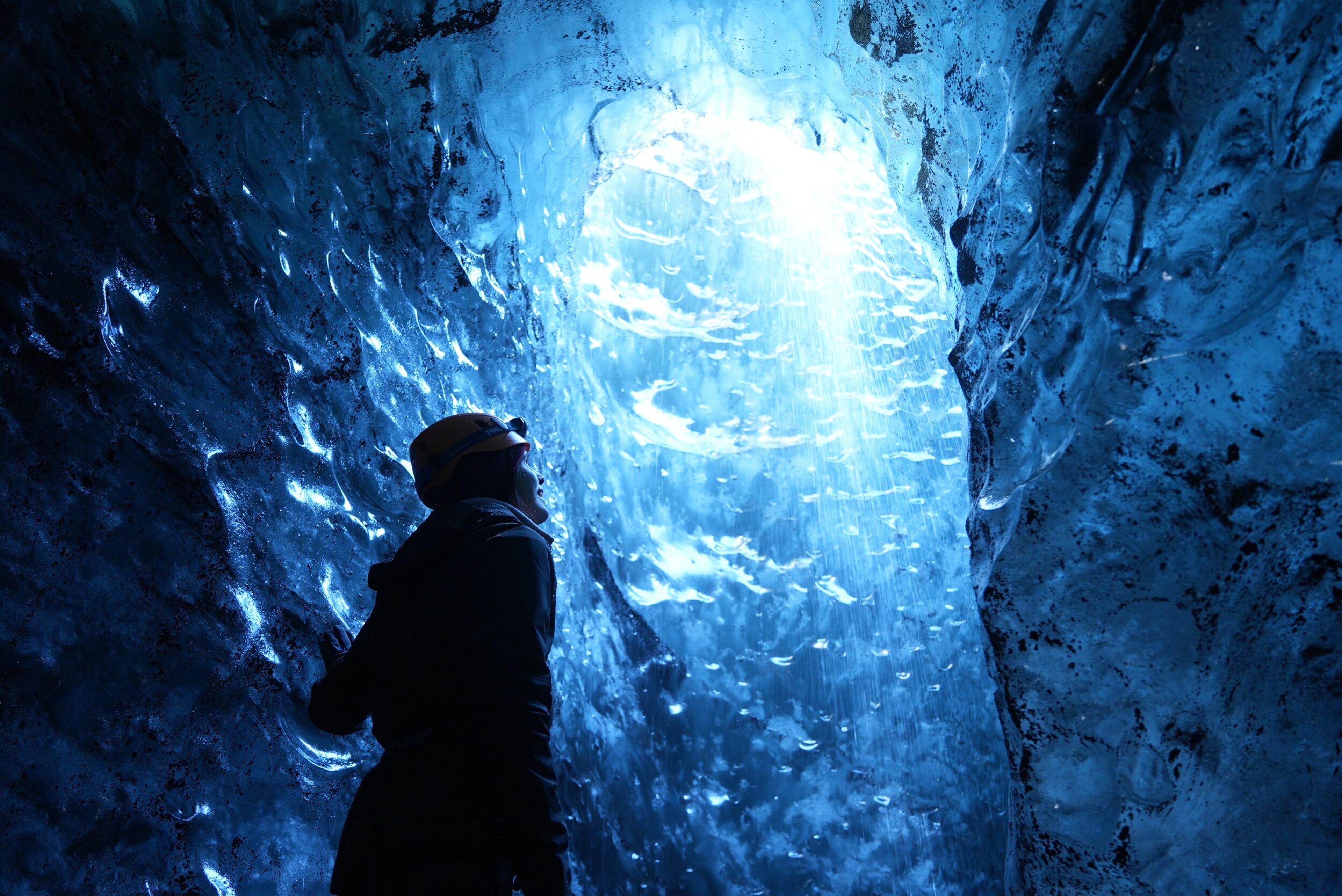 A person with a helmet standing in a blue ice cave, looking up at the light shining through the ice