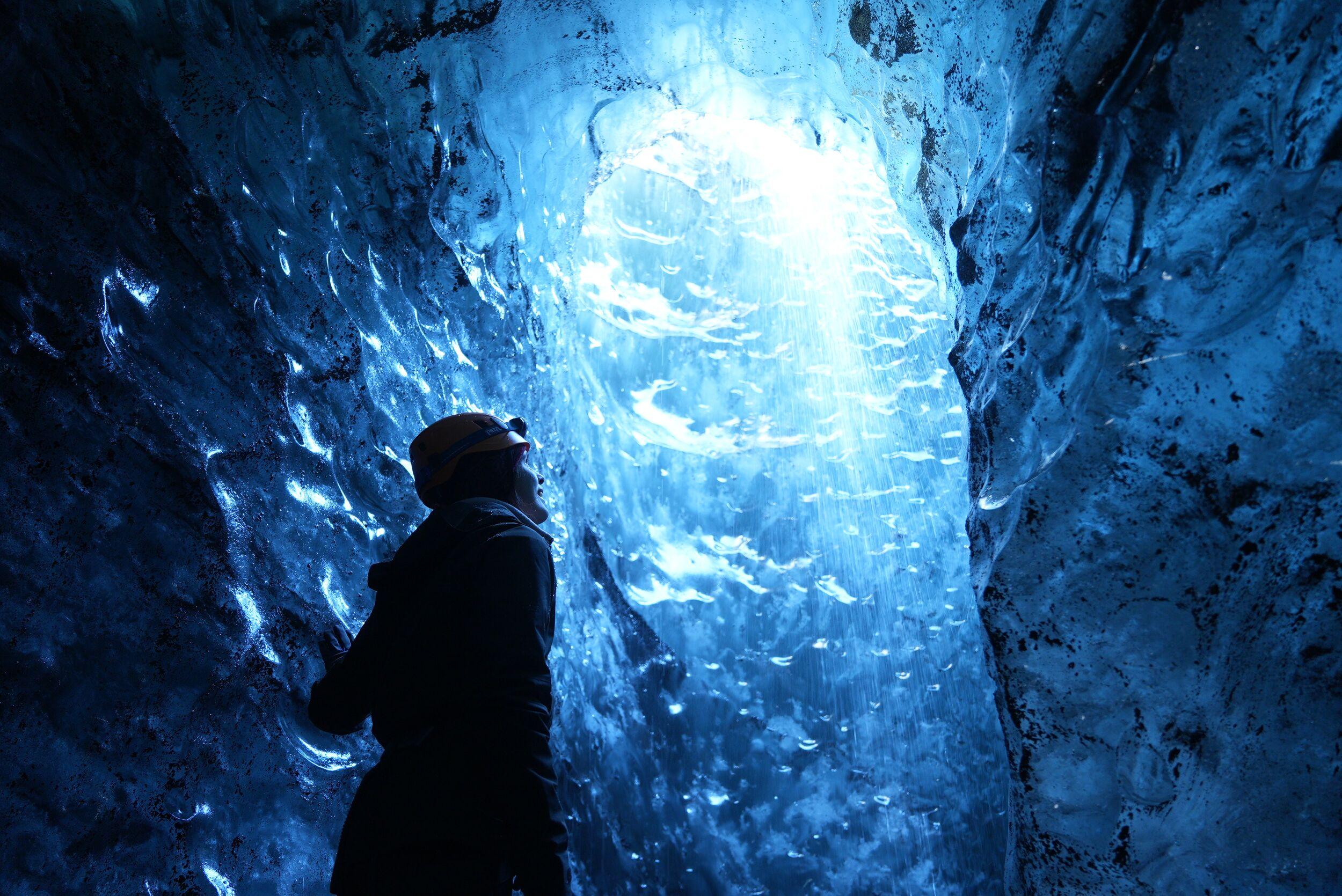 A person with a helmet standing in a blue ice cave, looking up at the light shining through the ice