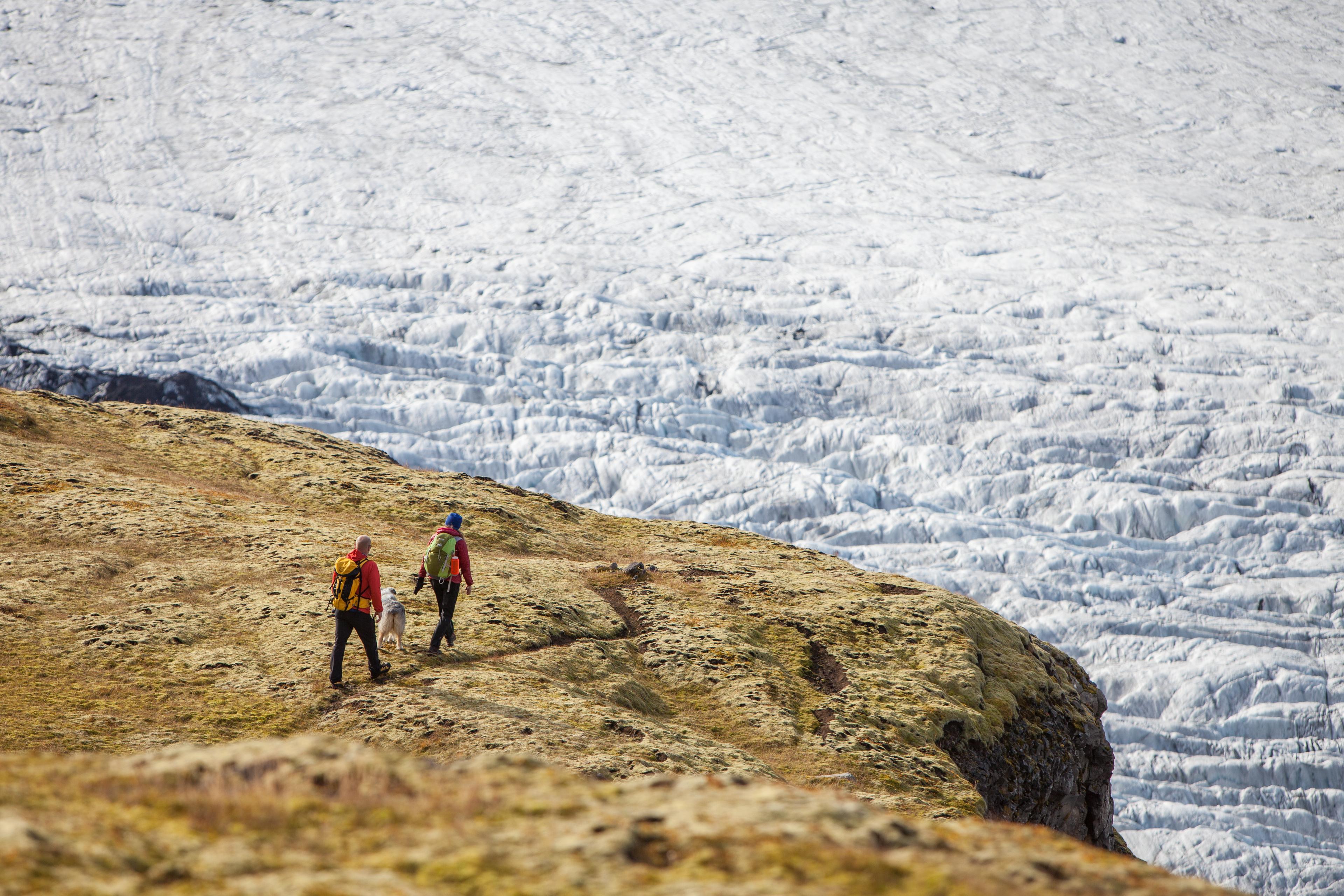 Two hikers getting close to the edge of a rock and looking down on the glacier below