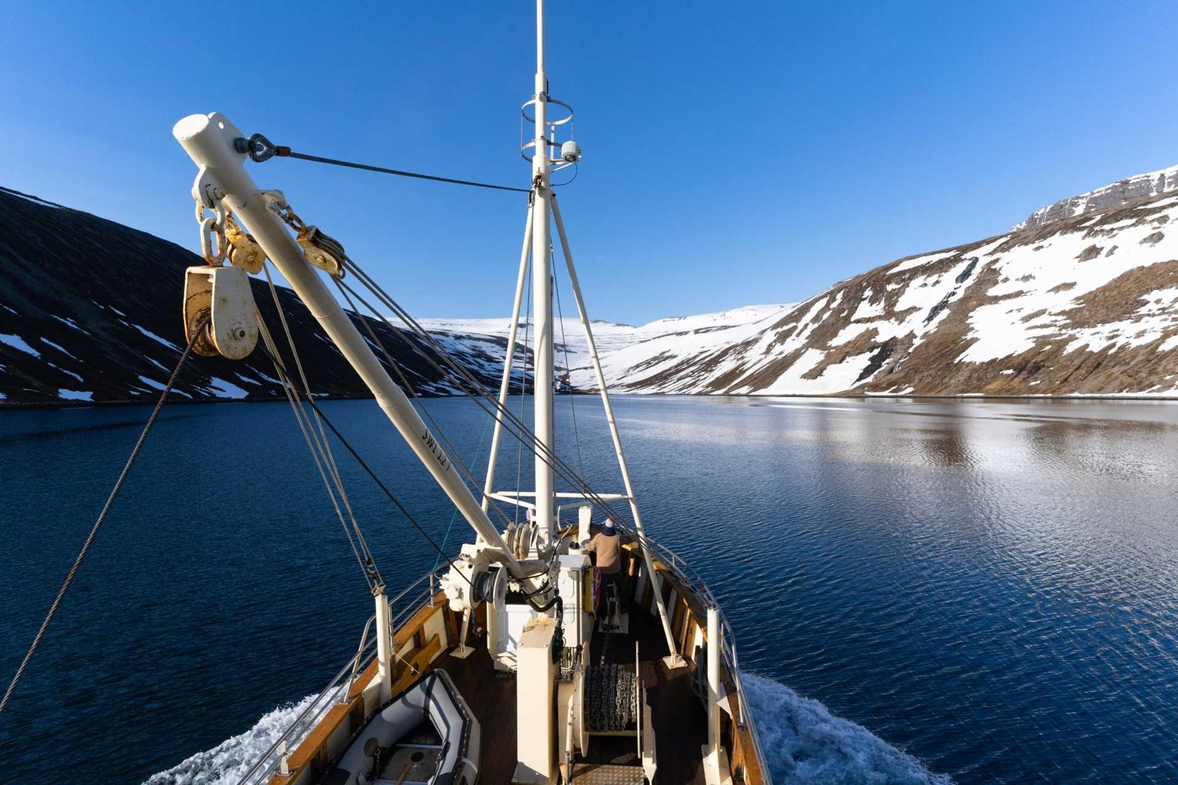 The bow of a ship cuts through calm blue waters, surrounded by snow-capped hills under a clear sky.