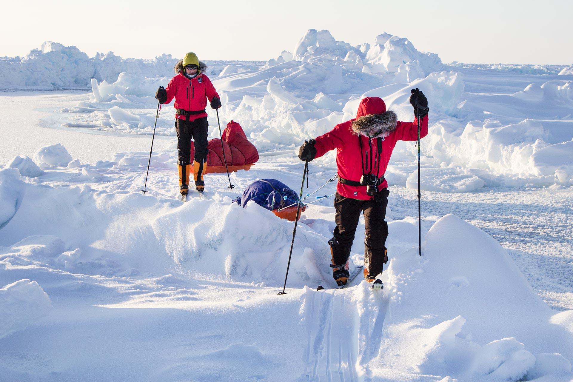 Two participants in a polar training course trek through deep snow, pulling sleds loaded with gear across a frozen landscape under bright sunlight.