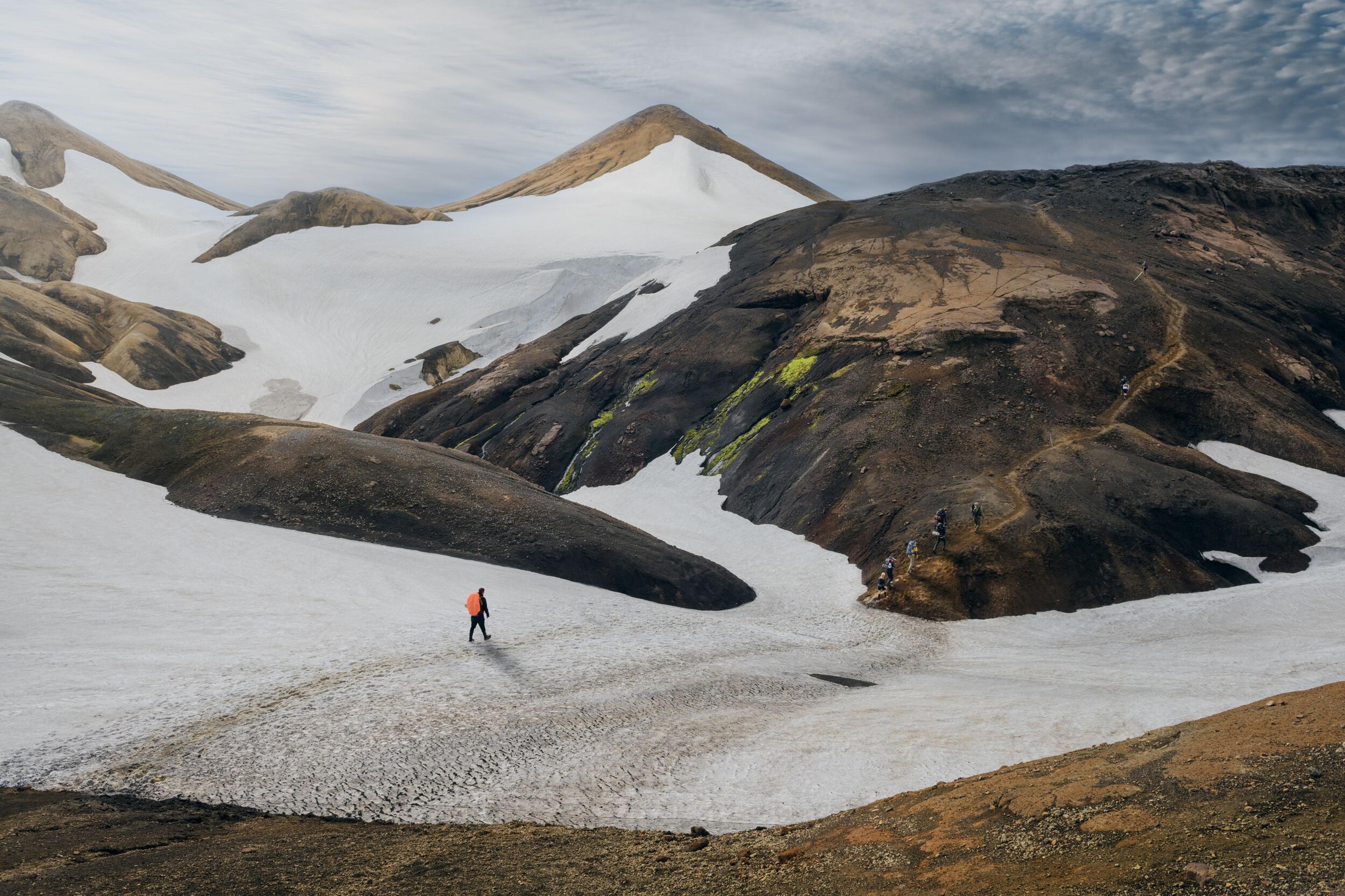 A hiker in an orange jacket treks across a snowy path surrounded by rugged, volcanic mountains along the Laugavegur trail in Iceland.