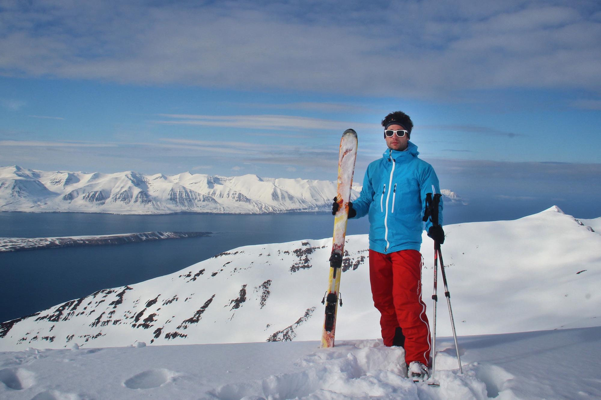 A man in blue posing in front of a snowy mountains and the ocean with his skis